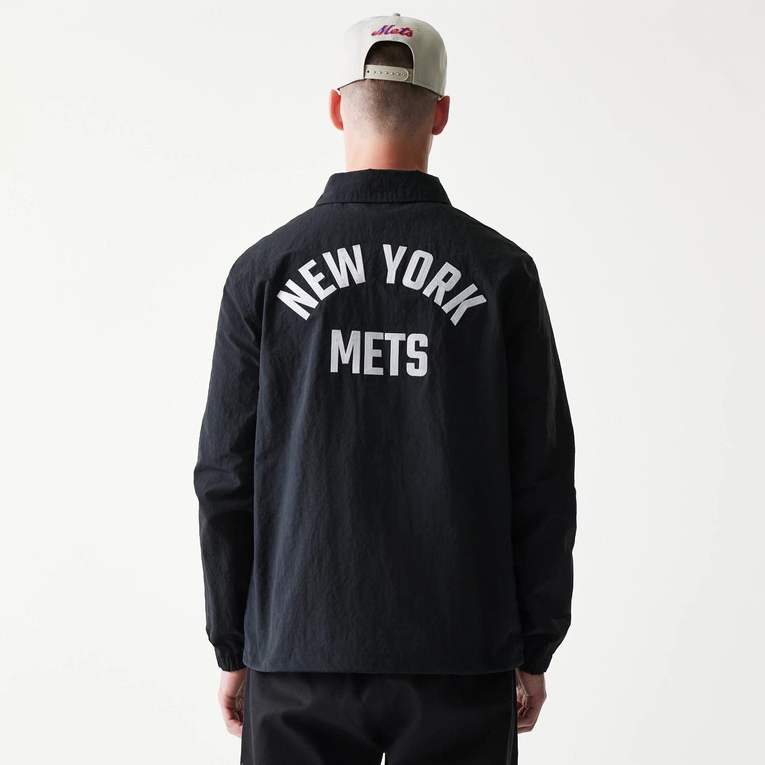 New Era New York Mets MLB Coaches Black Jacket, back view on a model with a light-coloured cap, featuring white New York Mets lettering across the upper back against the dark black fabric of the jacket.