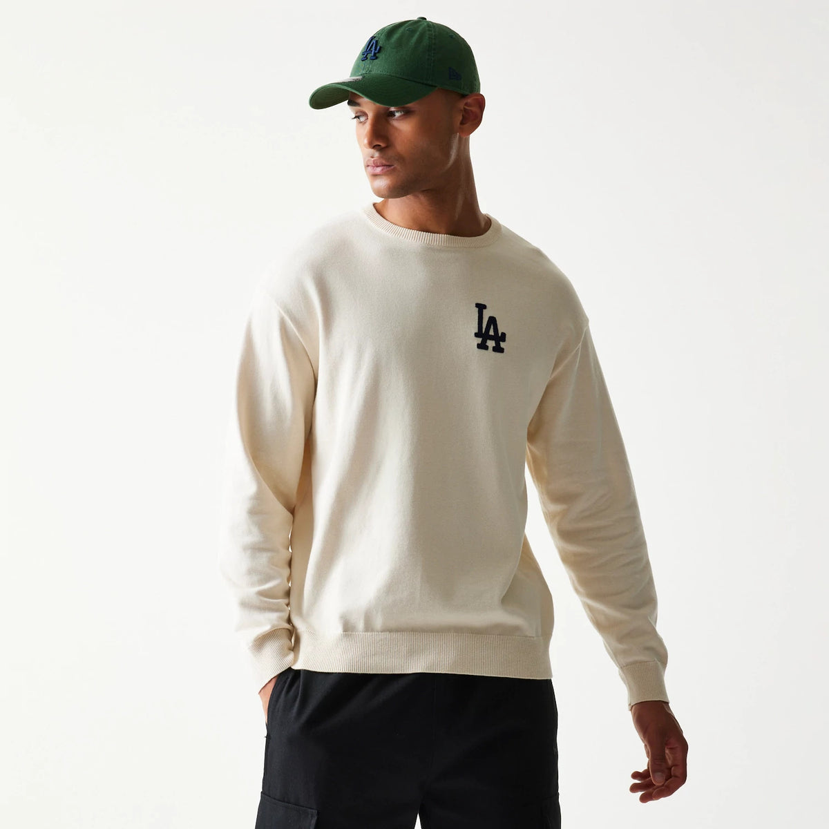 New Era LA Dodgers MLB Essential Open White Knitted Sweater, on-model front view showing the navy LA logo on the left chest, paired with a green cap, highlighting the sweater's relaxed fit and long sleeves.