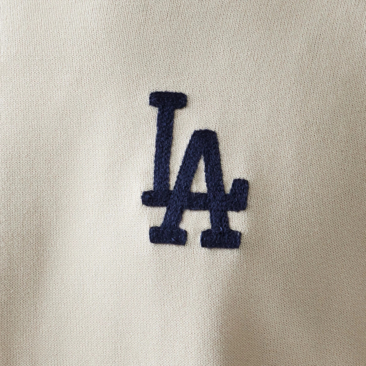 New Era LA Dodgers MLB Essential Open White Knitted Sweater, close-up showing the navy LA logo in raised embroidery on the soft white knit fabric, highlighting its texture and detail.
