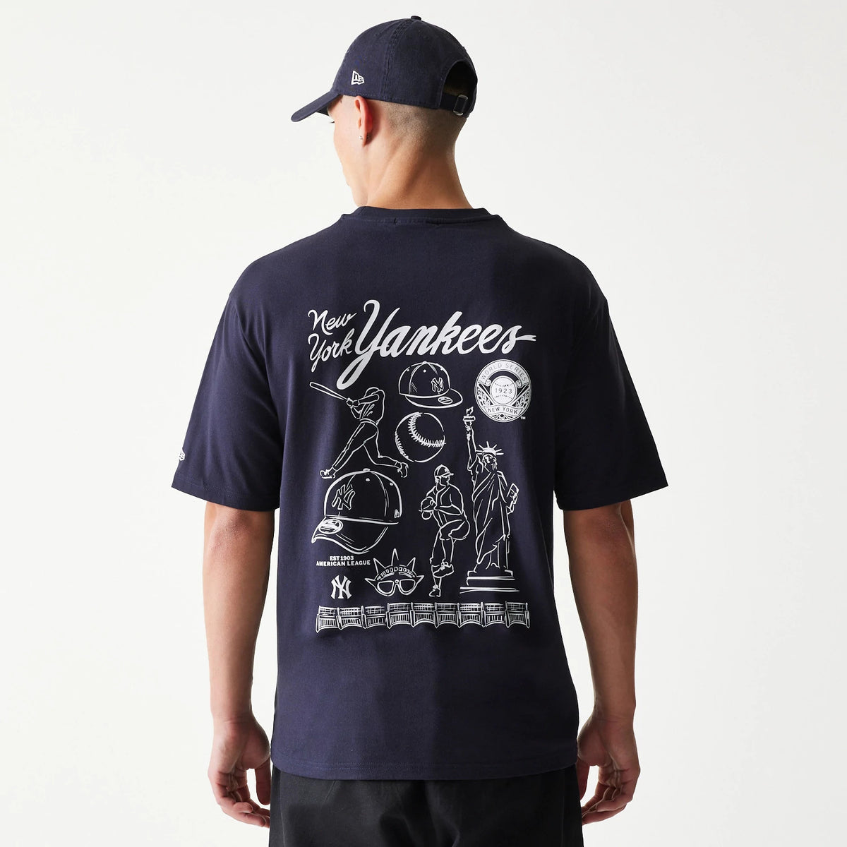 New Era New York Yankees MLB Baseball Graphic Navy Oversized T-Shirt, back view on a model featuring white graphic elements including baseball players, caps, a baseball, and Statue of Liberty.