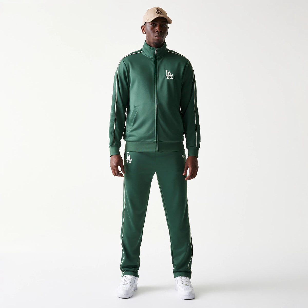 New Era LA Dodgers MLB Piping Dark Green Straight Leg Joggers, front view on a model with an LA Dodgers cap and matching jacket, showing the white embroidered LA logo on the left thigh and piping detail along the sides.