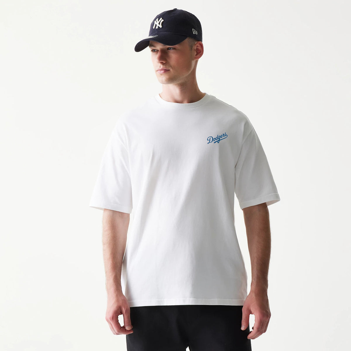 New Era LA Dodgers MLB World Series White T-Shirt, front view on-model showing the Dodgers logo embroidered in blue on the left chest, with the model styled in a contrasting black cap featuring a New York Yankees logo.