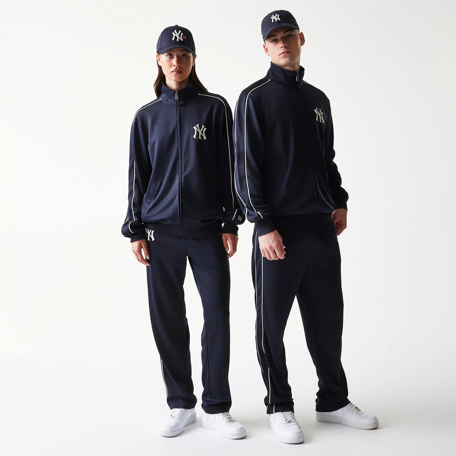 New Era New York Yankees MLB Piping Navy Track Jacket, front view on models wearing matching caps, showing the white embroidered New York Yankees logo on the left chest and contrasting piping running down the sleeves and sides.