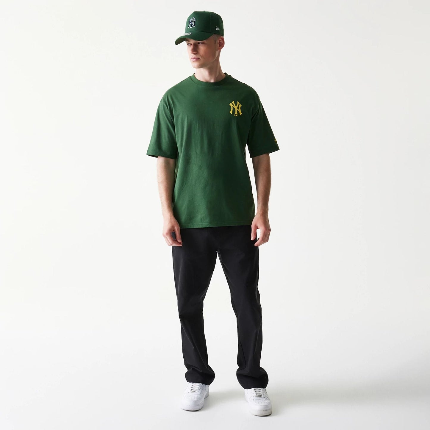 New Era New York Yankees MLB World Series Dark Green T-Shirt, front view on a model, featuring a yellow embroidered Yankees logo on the chest and styled with a matching dark green New Era cap and black trousers.