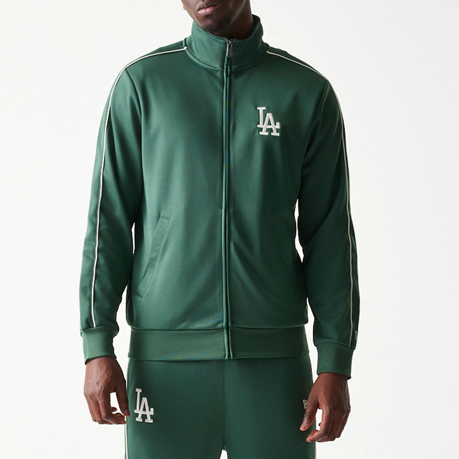 New Era LA Dodgers MLB Piping Dark Green Track Jacket, front view on-model showing the white embroidered LA logo on the left chest, a high collar with a full zip, and white piping detail along the shoulder and sleeves.