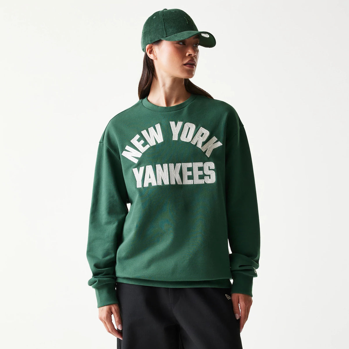 New Era New York Yankees MLB Graphic Dark Green Oversized Crew Neck Sweater, front view on a model with large white lettering across the chest, long sleeves and ribbed neckline, paired with black trousers and a matching dark green cap.