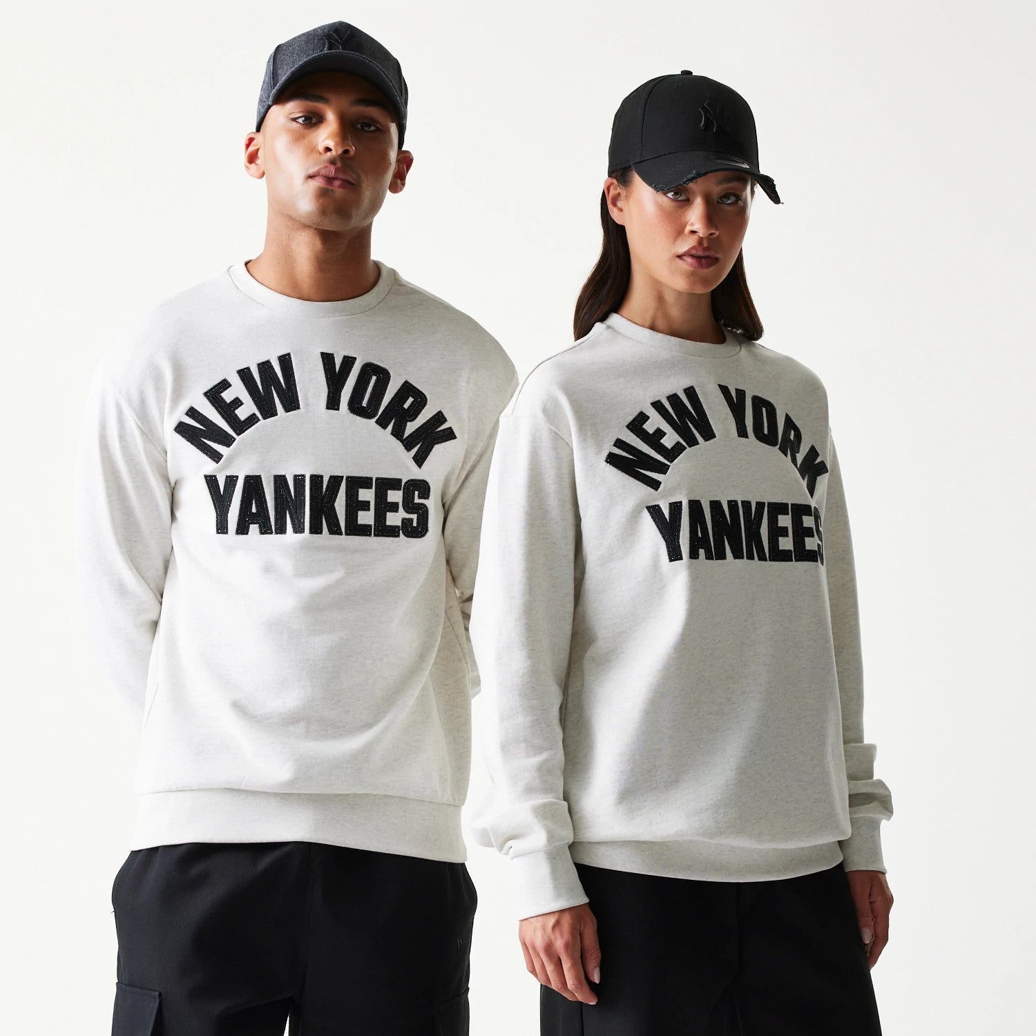 New Era New York Yankees MLB Graphic Pastel Grey Oversized Crew Neck Sweater, front view on models with matching black caps, featuring black New York Yankees lettering across the chest and ribbed cuffs at the sleeves.