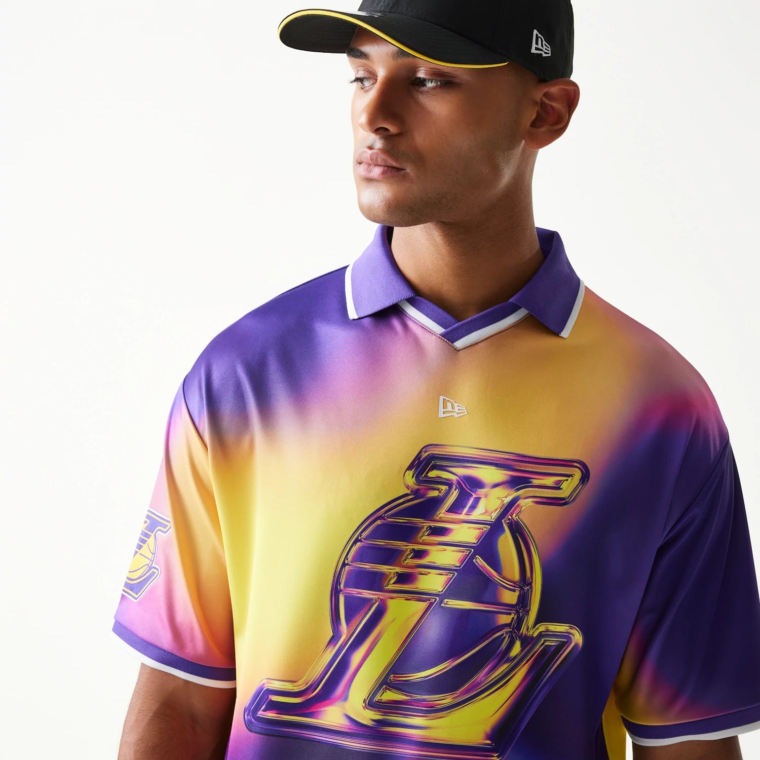 New Era LA Lakers NBA All Over Print Purple T-Shirt, front three-quarter view on a model with a cap showing a large, colourful LA Lakers graphic across the chest, purple and yellow gradient design, and an embroidered New Era logo beneath the collar.