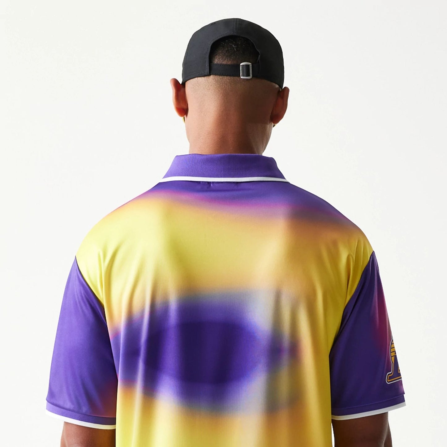 New Era LA Lakers NBA All Over Print Purple T-Shirt, back view on a model paired with a black cap, showing a gradient design with purple and yellow tones, accented by a white trim on the collar and sleeves.