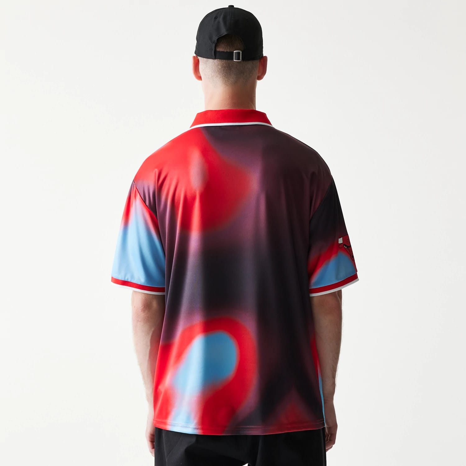 New Era Chicago Bulls NBA All Over Print Red T-Shirt, back view on a model with a black cap, showing a red and blue abstract pattern covering the entire shirt and an embroidered Bulls logo on the sleeve against a dark gradient background.