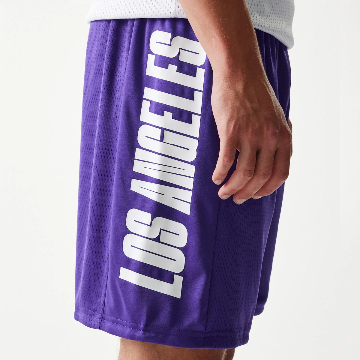 New Era LA Lakers NBA Mesh Purple Shorts, side view showing white Los Angeles lettering running vertically down the side of the purple shorts, worn slightly above the knee by a model with a white top.