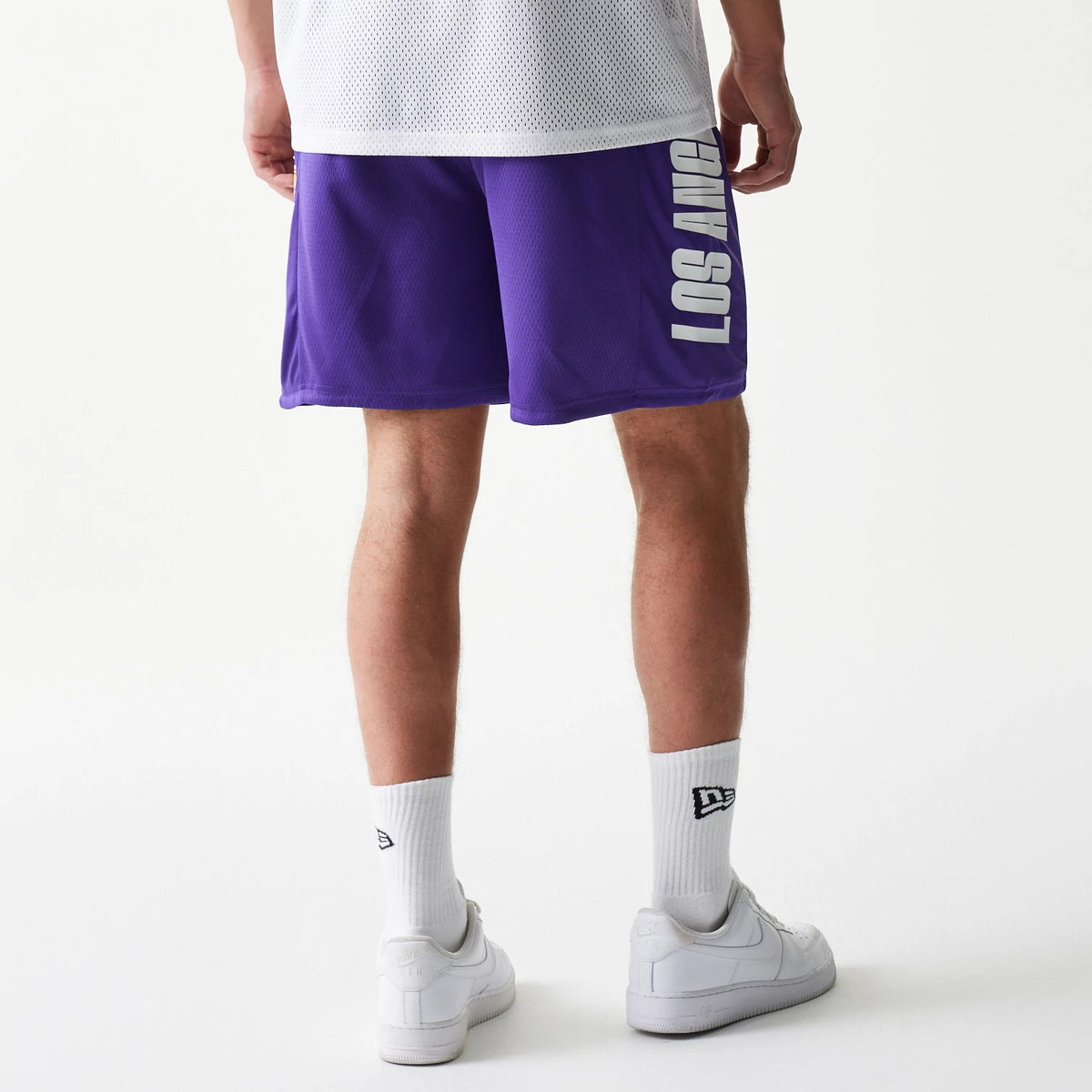 New Era LA Lakers NBA Mesh Purple Shorts, back view showing the purple colour with white LOS ANGELES lettering in down the side, styled casually on a model paired with a white mesh top and trainers.
