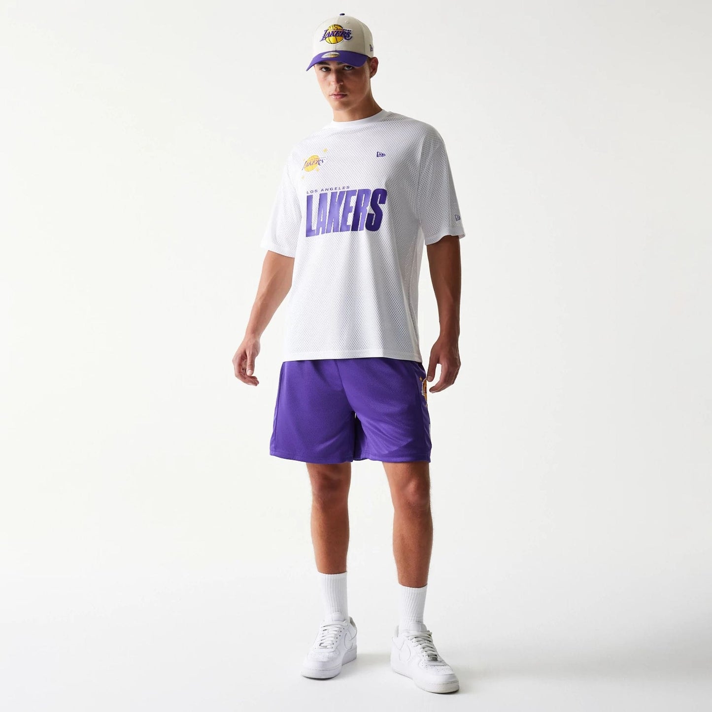New Era LA Lakers NBA Mesh Purple Shorts, front view on-model with a white Lakers T-shirt and cap, showing the rich purple colour of the shorts with the Lakers logo prominently displayed on the left leg.