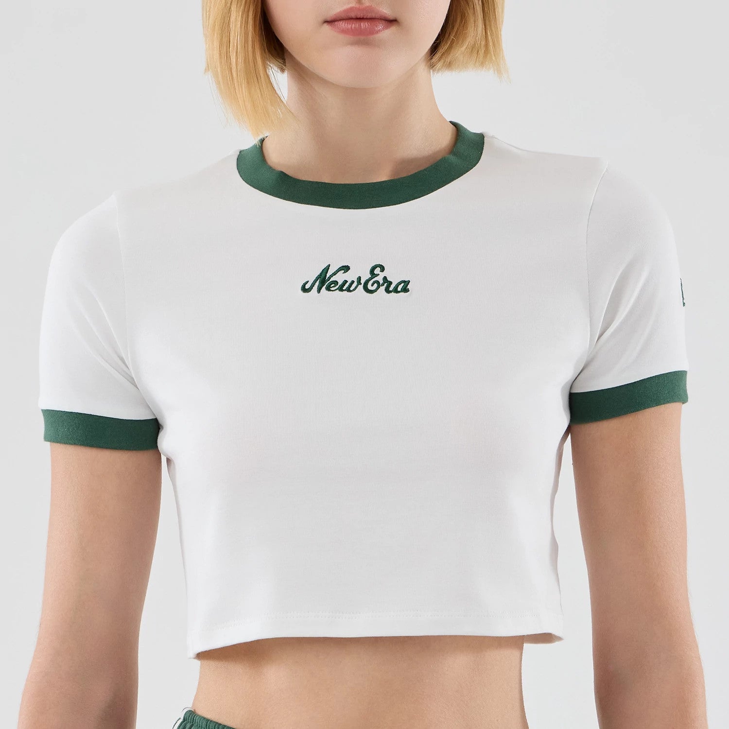 Womens New Era White Cropped Slim T-shirt, front view on-model showing a white cropped tee with green embroidered New Era logo on the chest, green ribbed neckline and sleeve cuffs, paired with matching green shorts.