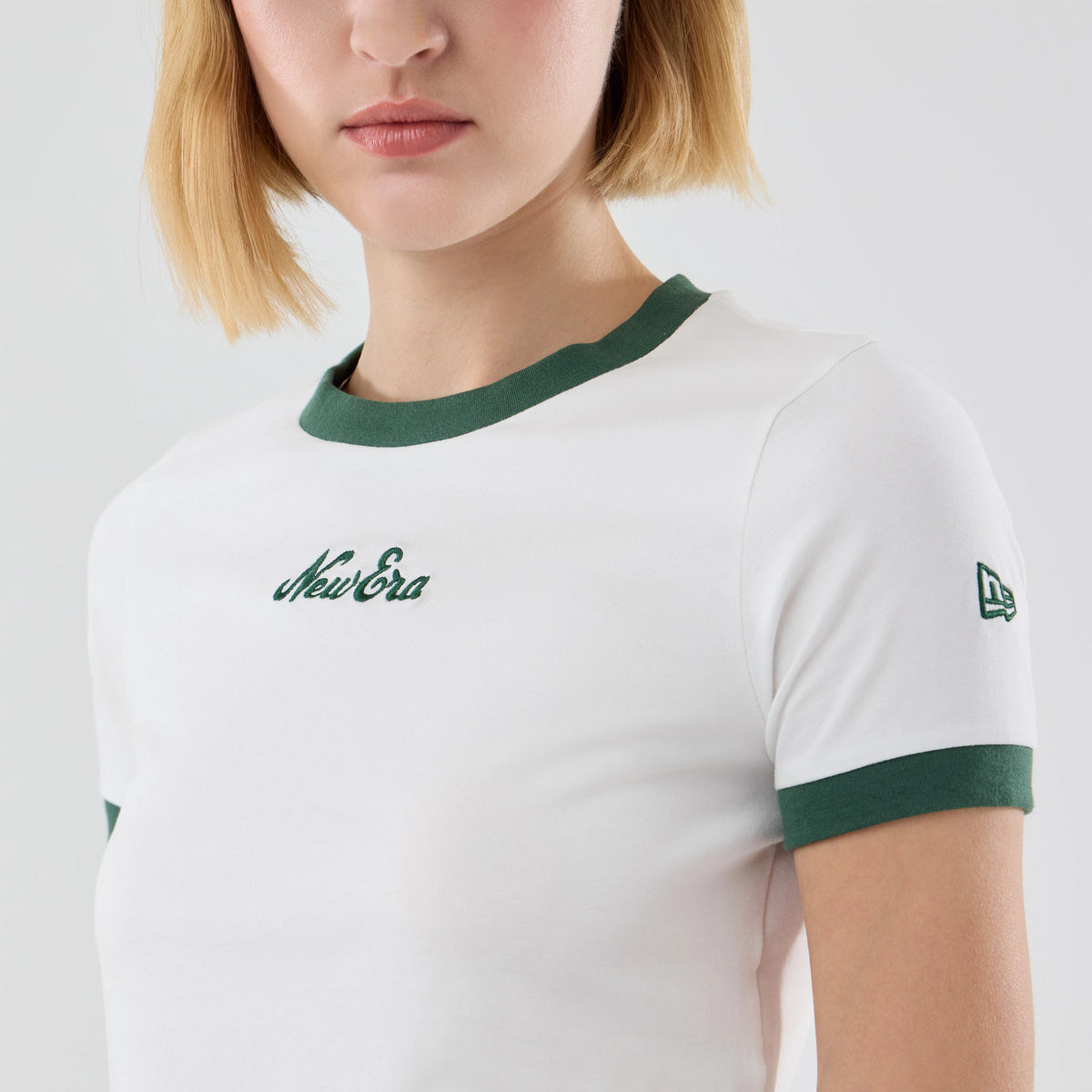 Womens New Era White Cropped Slim T-shirt, front view on-model showing the embroidered New Era script logo in green on the chest, with green trim on the collar and sleeves, and an embroidered New Era flag logo on the left sleeve.