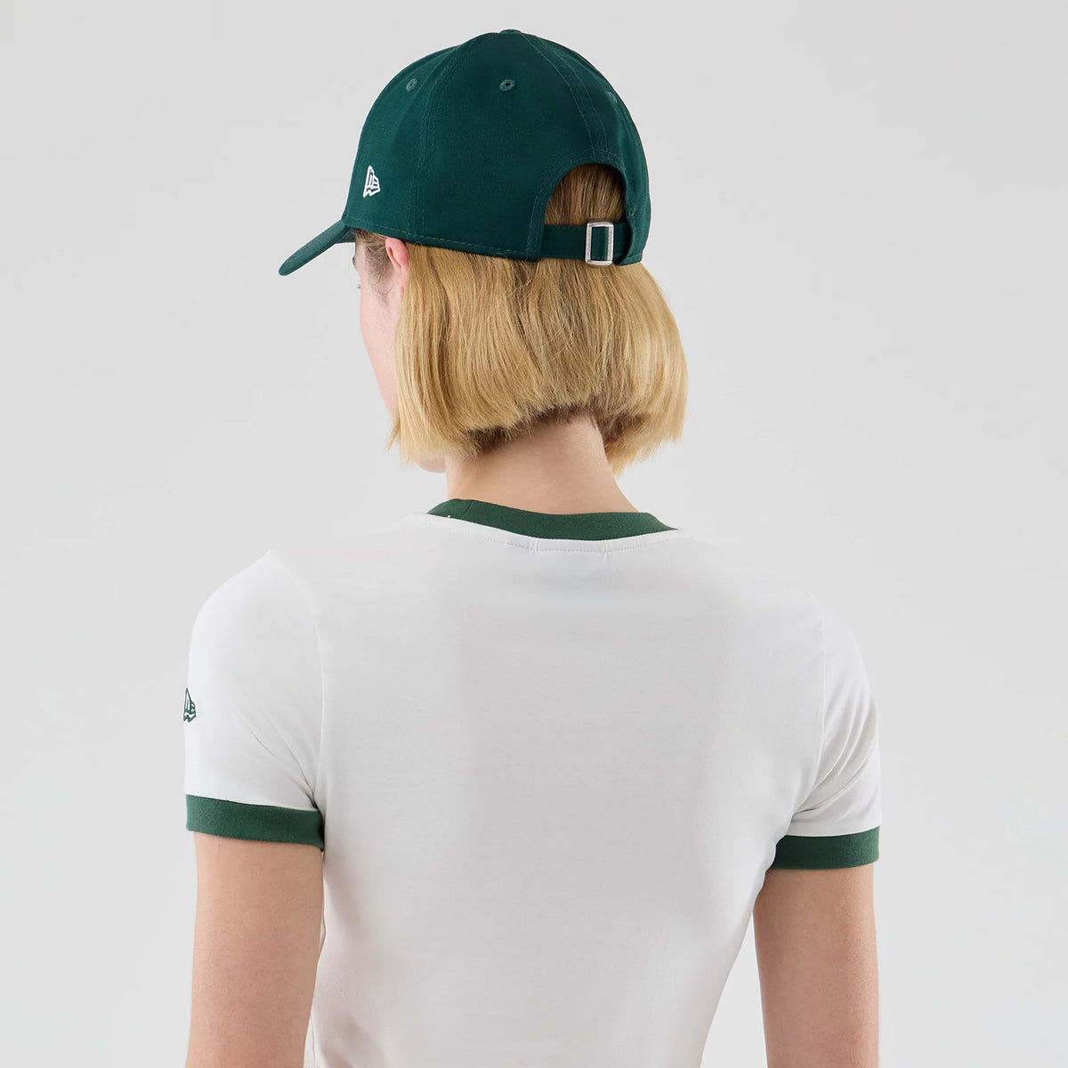 Womens New Era White Cropped Slim T-shirt, back view on a model pairing it with a green cap, highlighting the green collar and sleeve trim, and an embroidered New Era logo on the left sleeve.