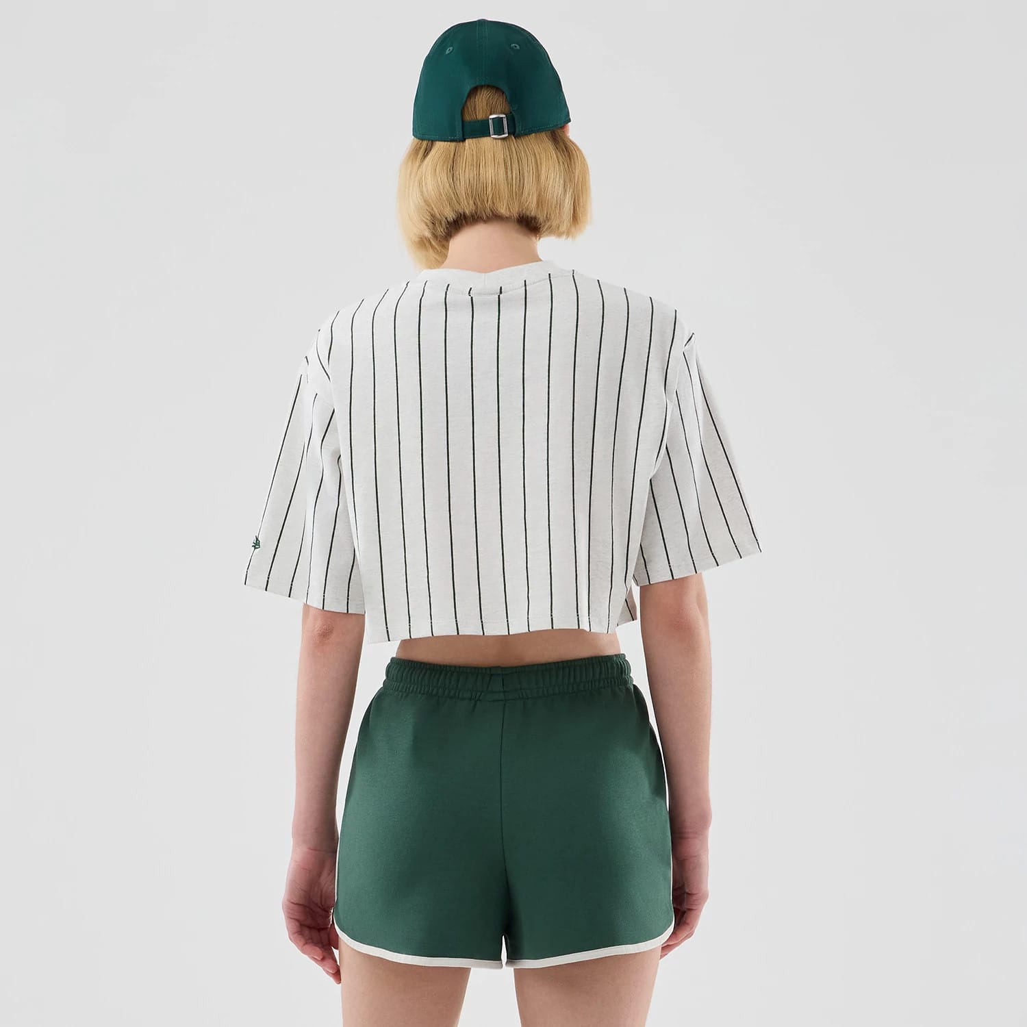Womens New Era Pinstripe Pastel Grey Cropped T-shirt, back view on a model with a matching green cap, featuring vertical black pinstripes across the light grey fabric and a small embroidered logo on the left sleeve.