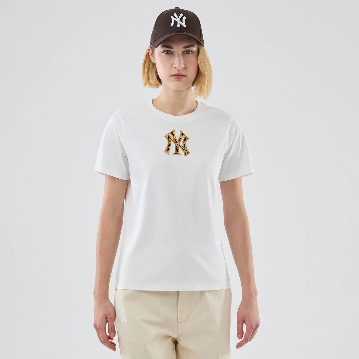 New Era New York Yankees Womens MLB Infill White T-shirt, front view on-model showing a patterned NY logo embroidered in gold and black in the centre of the chest, styled with beige trousers featuring a matching NY logo on the left leg.