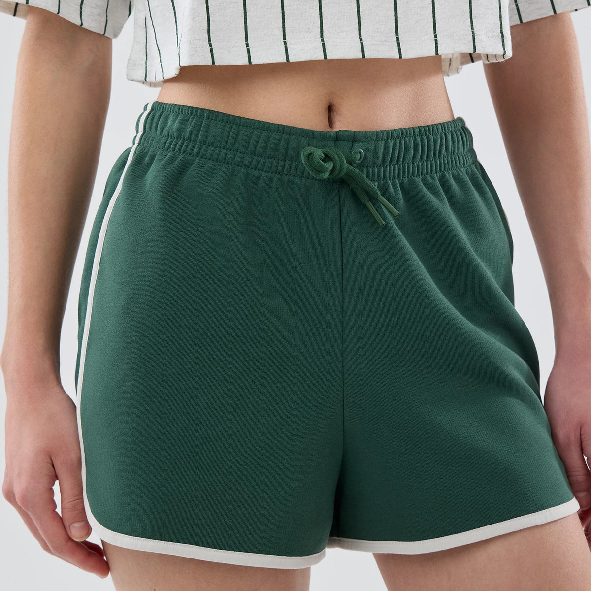 Womens New Era Dark Green Track Shorts, front view on-model showing dark green shorts with a drawstring waistband and white piping along the sides and hem, paired with a cropped striped top.
