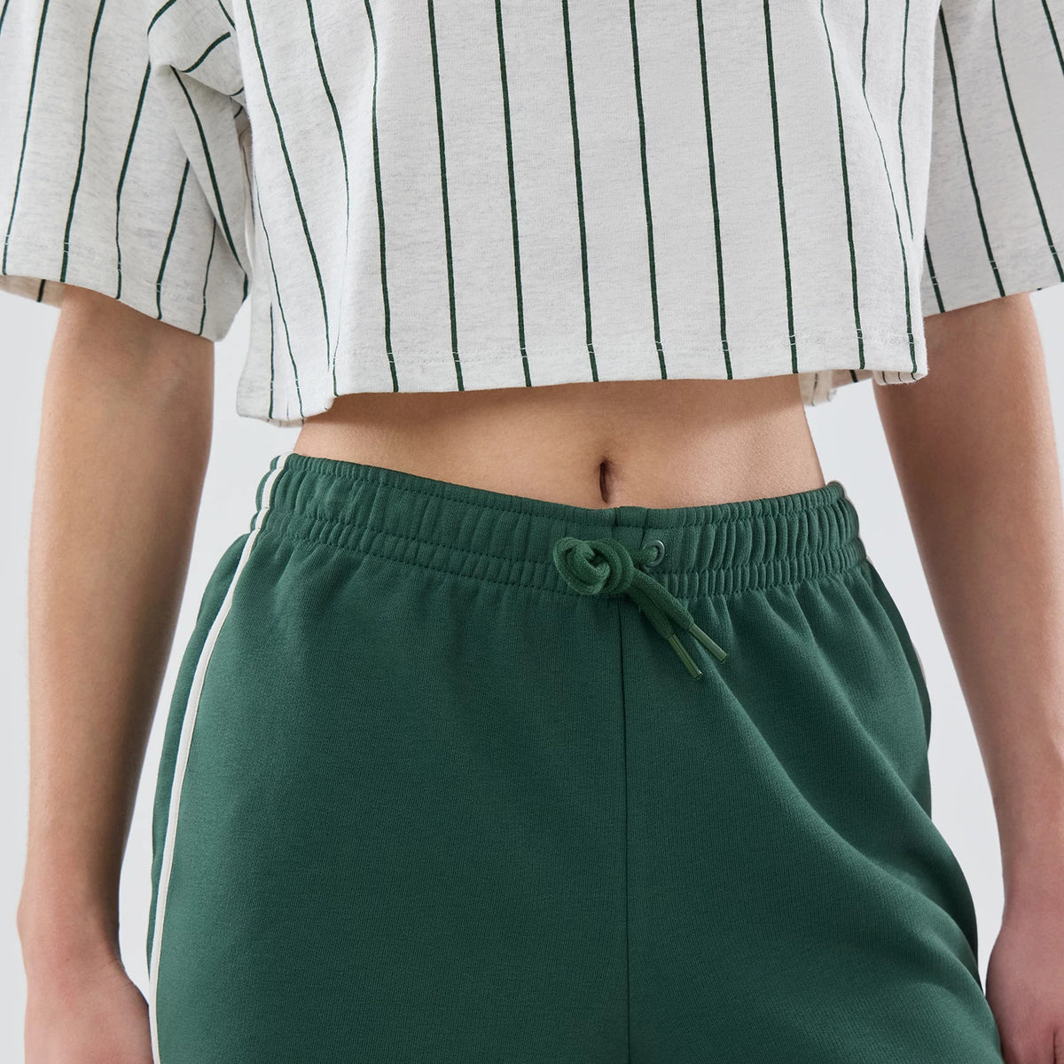 Womens New Era Dark Green Track Shorts, front view showing the elasticated waistband with drawstring adjustment, dark green colour with white side stripes, paired with a white pinstriped crop top on-model.