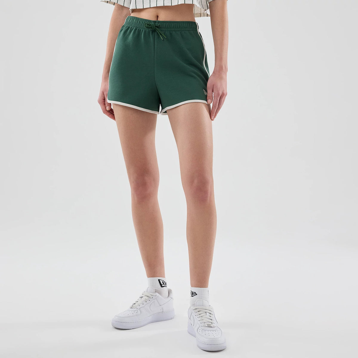 Womens New Era Dark Green Track Shorts, front view on a model wearing a cropped striped top and white trainers, featuring white trim on the hem and a embroidered New Era logo near the hemline.