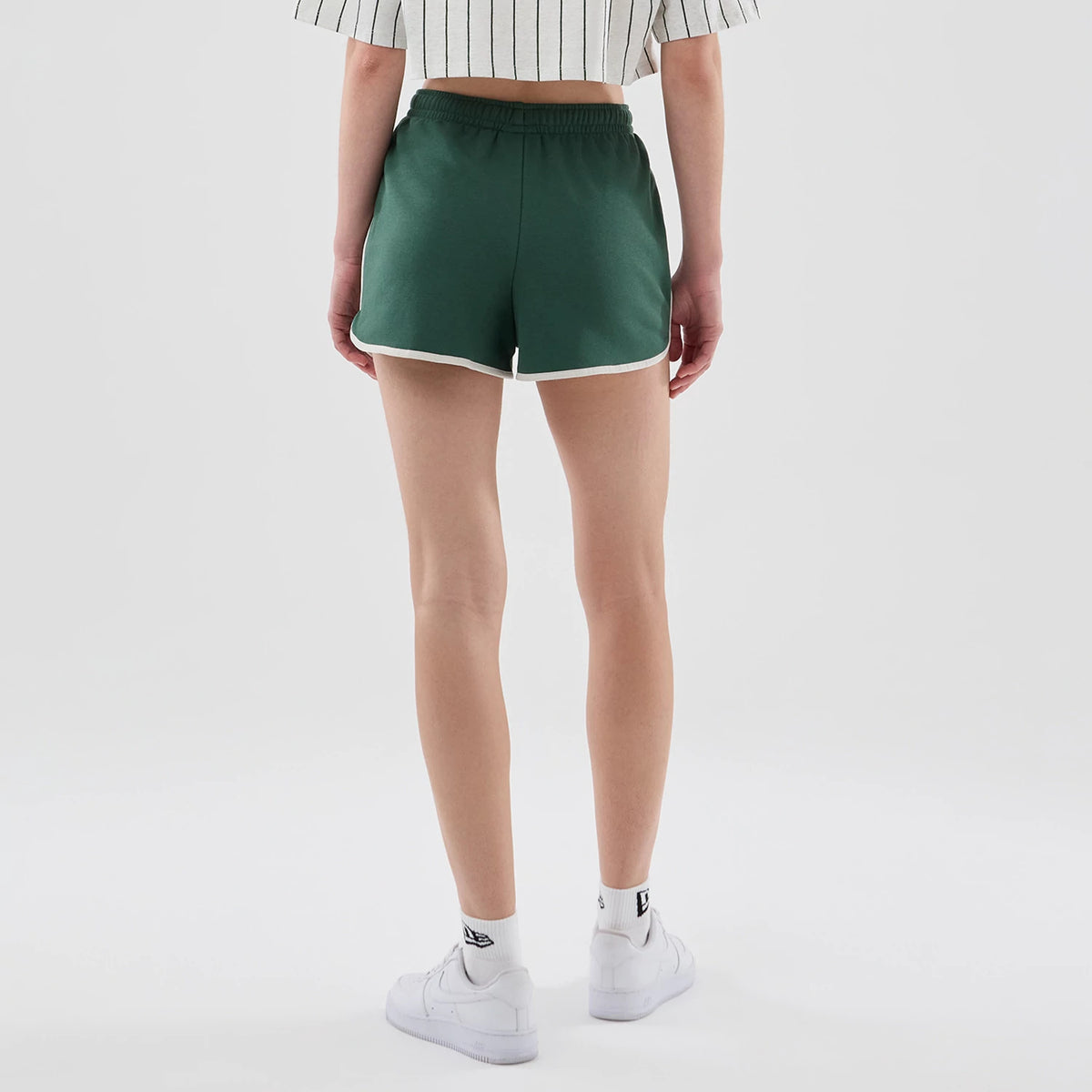 Womens New Era Dark Green Track Shorts, back view on-model showing dark green fabric with white trim along the hem and an elastic waistband, styled with a short-sleeve top and white trainers.