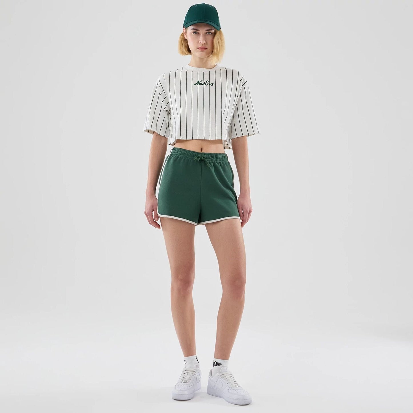 Womens New Era Dark Green Track Shorts, front view on-model showing the dark green shorts with white trim and a drawstring waist, paired with a striped crop top featuring an embroidered New Era logo and a matching dark green cap.