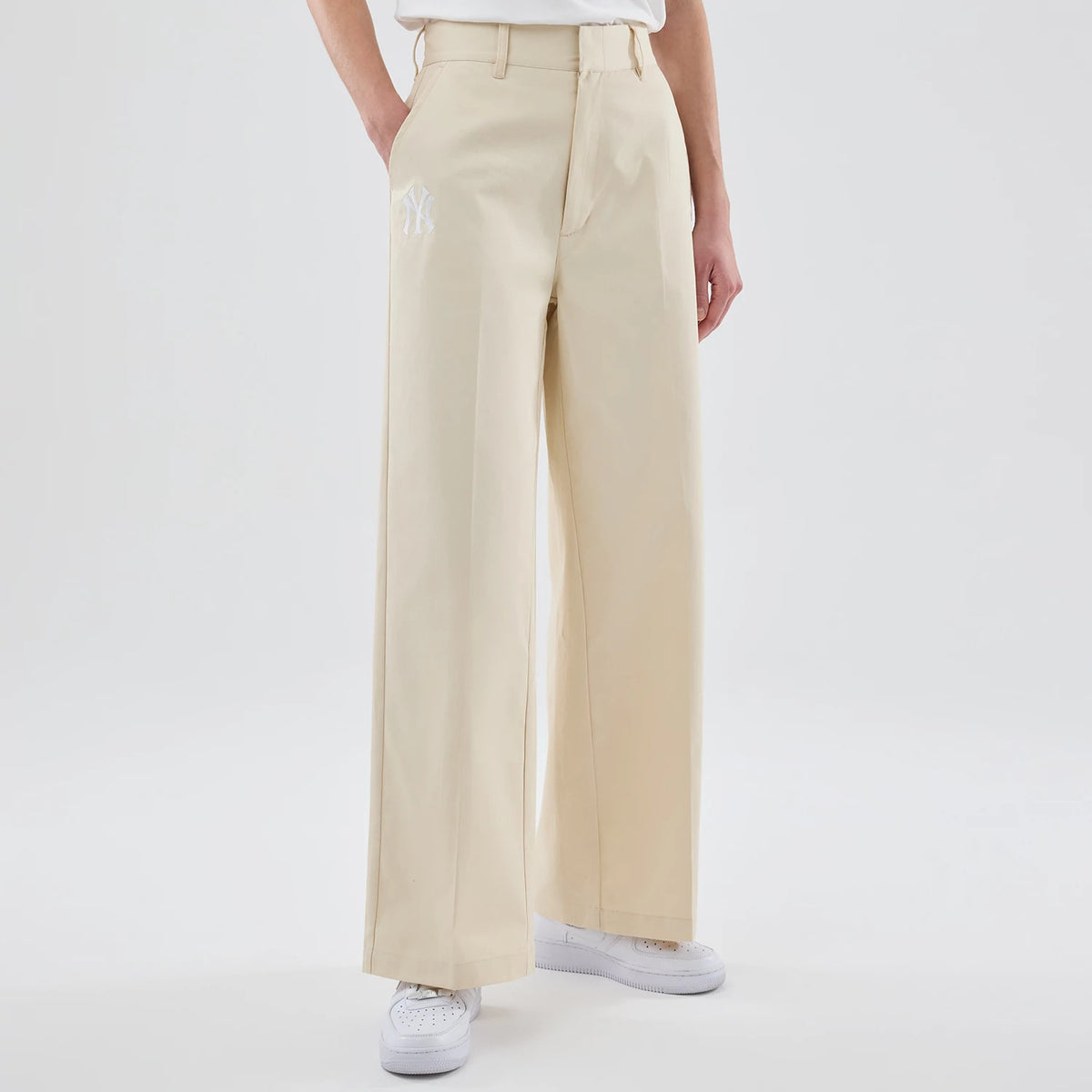 New Era New York Yankees Womens MLB Cream Chino Trousers, front view on a model showing the white embroidered NY logo on the left thigh, with belt loops on the waistband and wide-leg fit paired with white trainers.