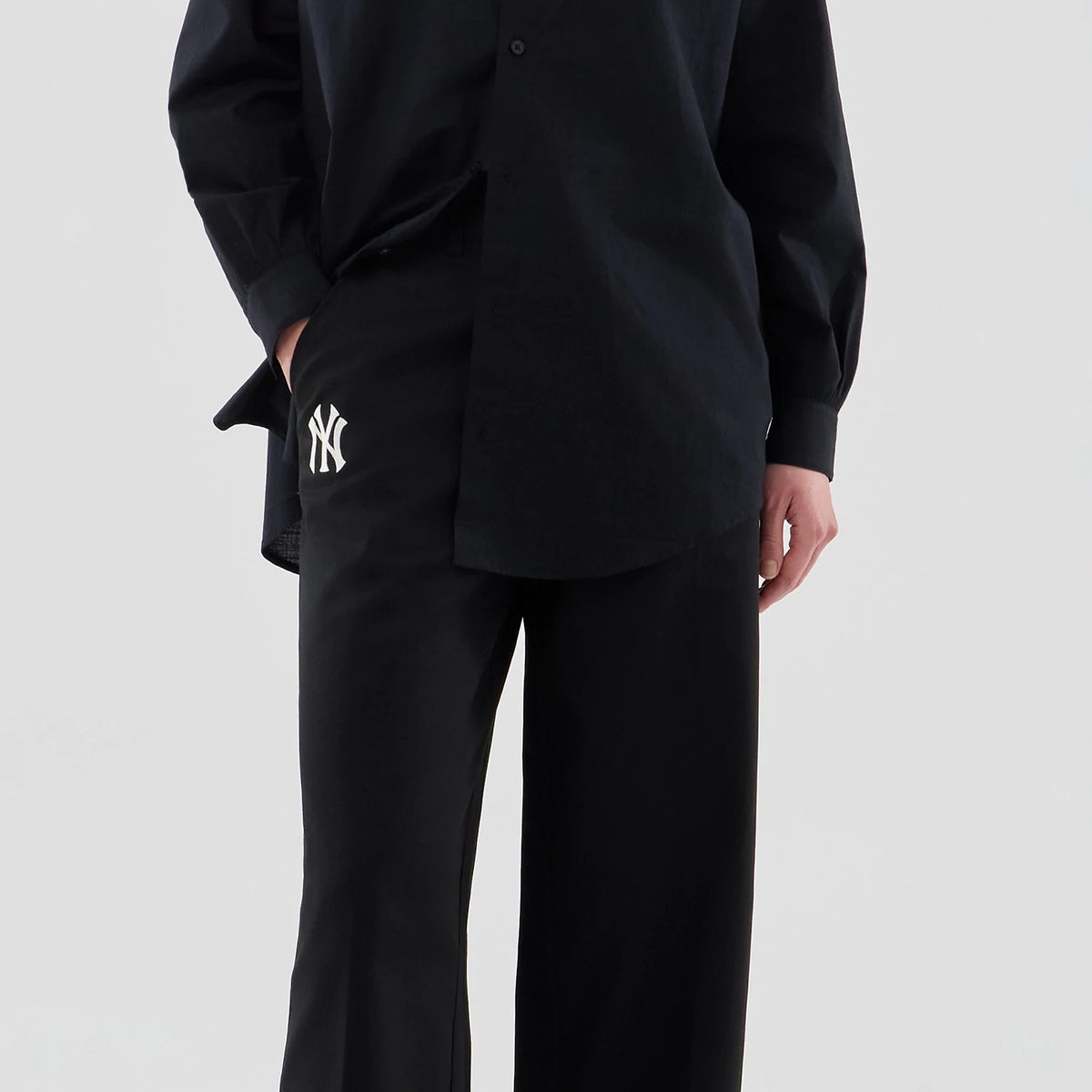 New Era New York Yankees Womens MLB Black Chino Trousers, front view on a model showing the white embroidered NY logo on the right pocket, with a relaxed fit and black buttoned shirt pairing.