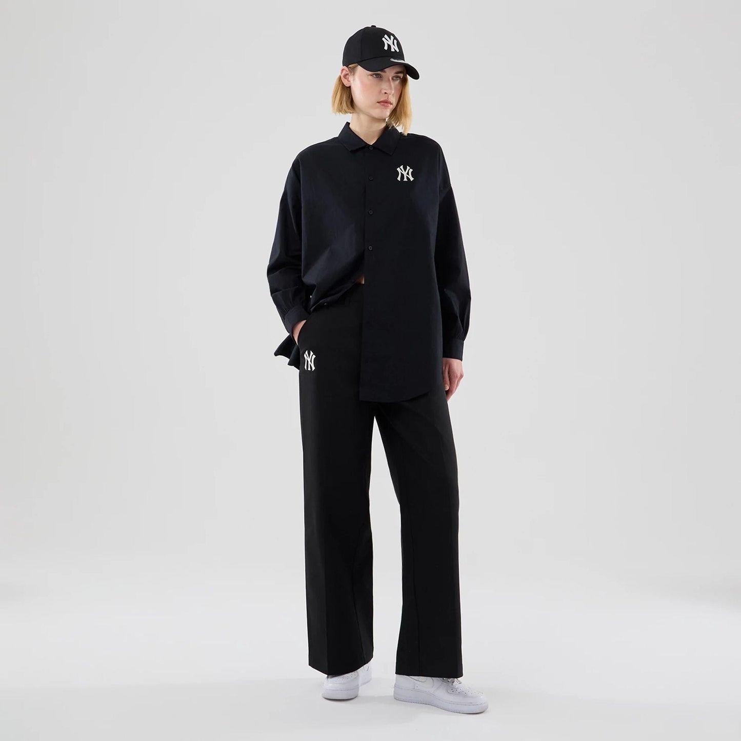 New Era New York Yankees Womens MLB Black Chino Trousers, on-model view showing the white embroidered NY logo on the right trouser pocket, paired with a matching oversized shirt and cap, highlighting a coordinated outfit.