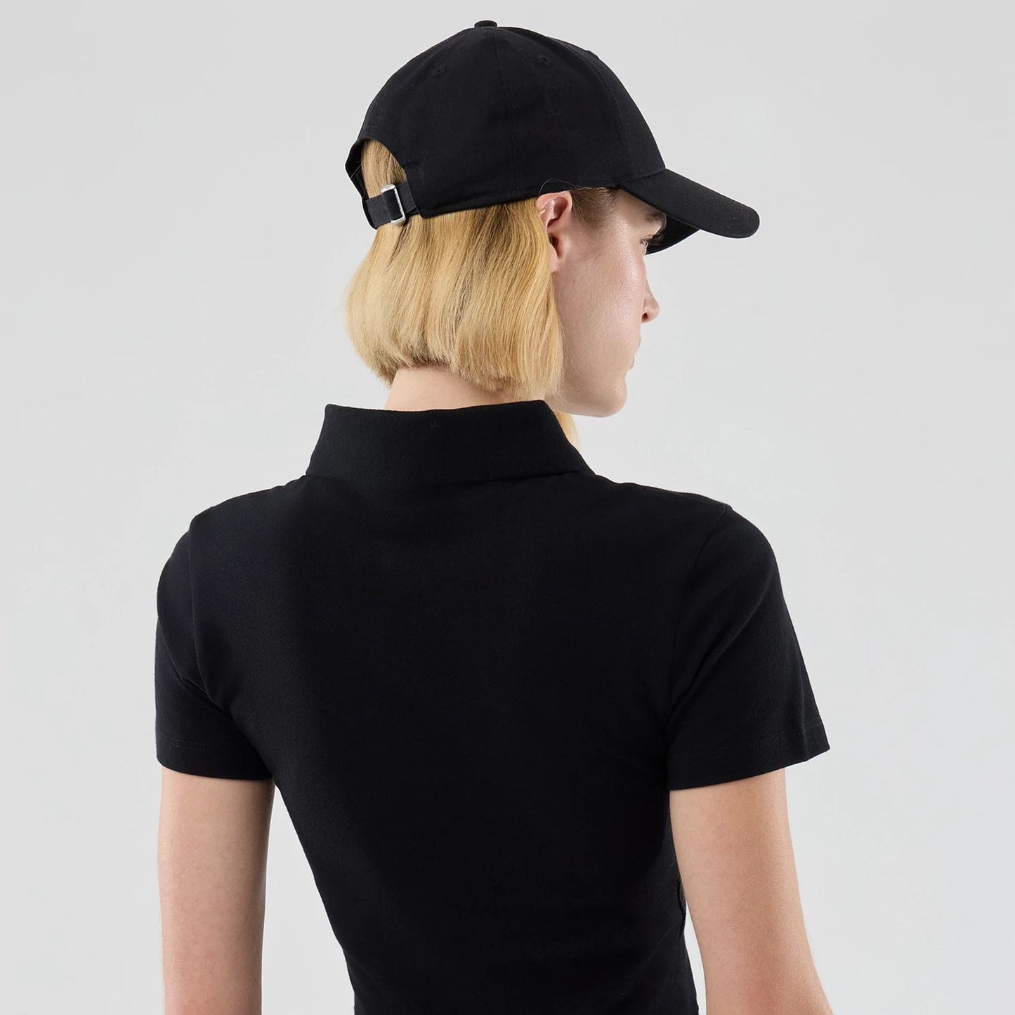 New Era New York Yankees Womens MLB Black Cropped Slim Polo T-shirt, back three-quarter view on-model showing the black fabric with a folded collar, short sleeves, and styled with a matching black cap featuring a buckle at the back.