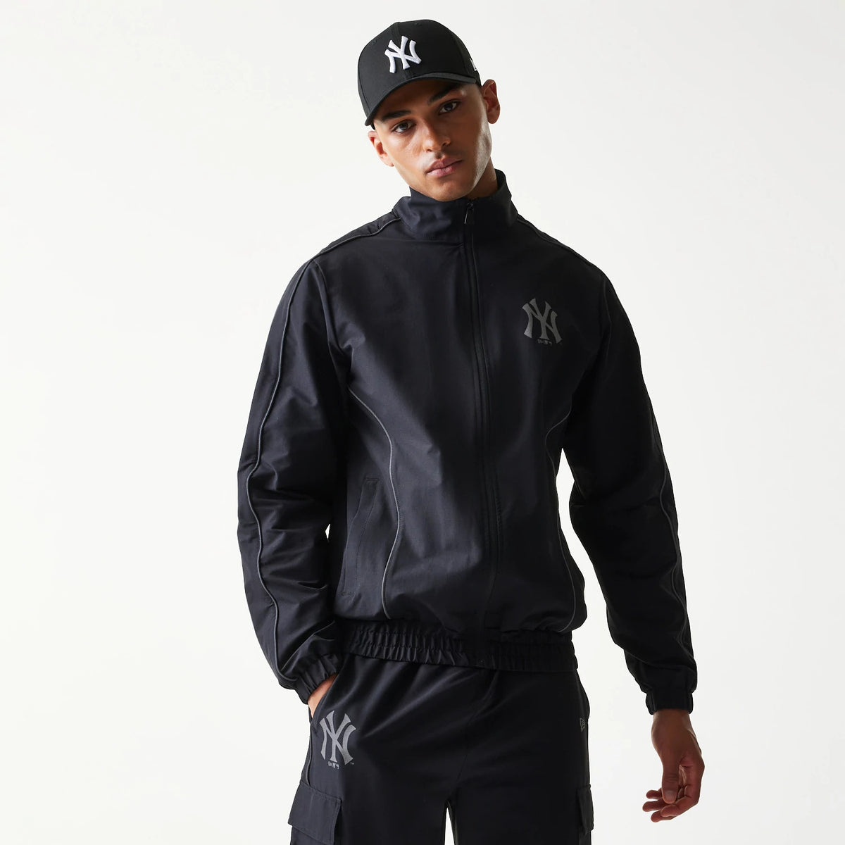 New Era New York Yankees MLB Black Track Jacket, front view on-model featuring the raised New York Yankees logo on the left chest, full zip closure, and matching black trousers with a logo near the pocket, styled with a black cap.
