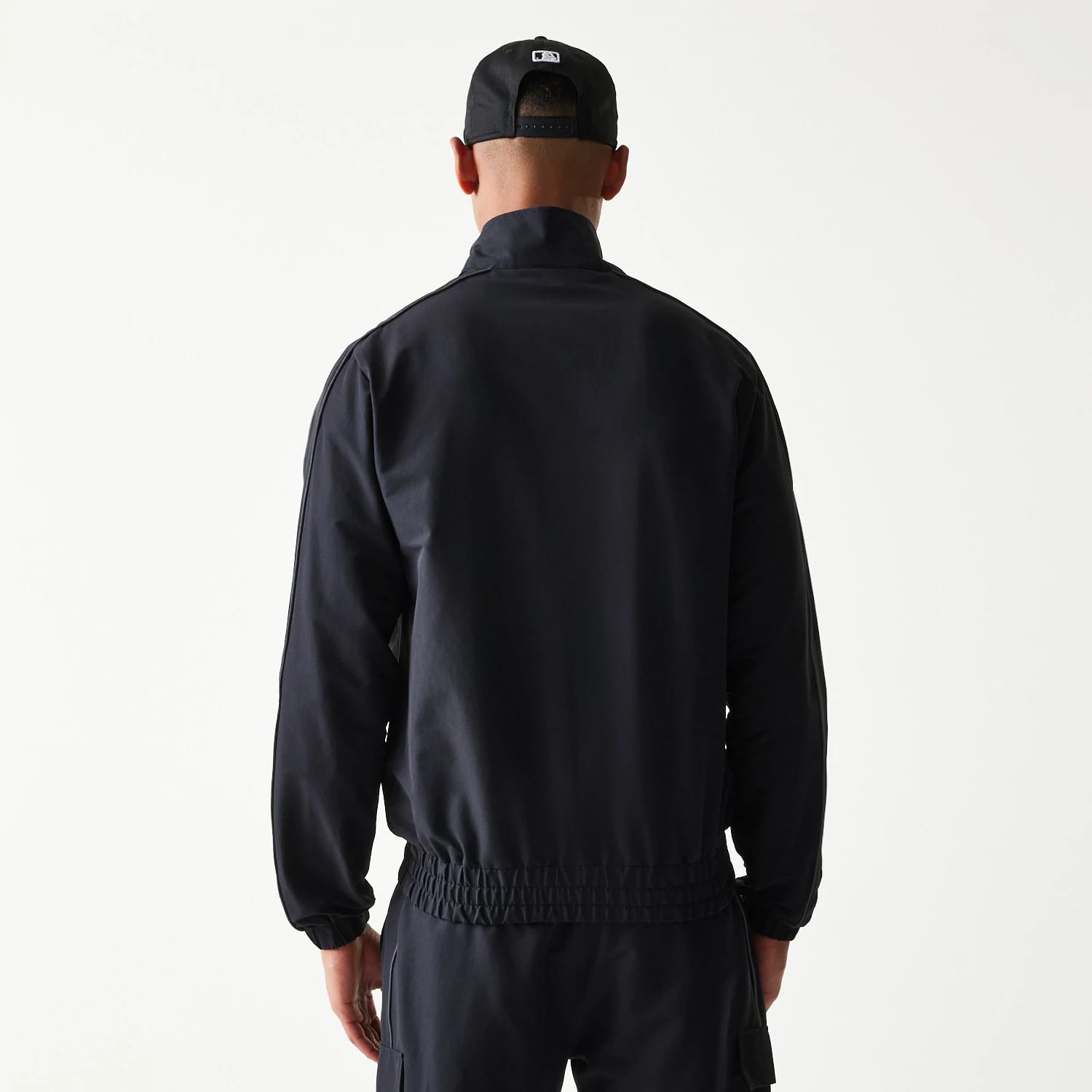 New Era New York Yankees MLB Black Track Jacket, back view on-model displaying the black back panel and an elasticated waist, paired with a black cap featuring an embroidered logo on the back.