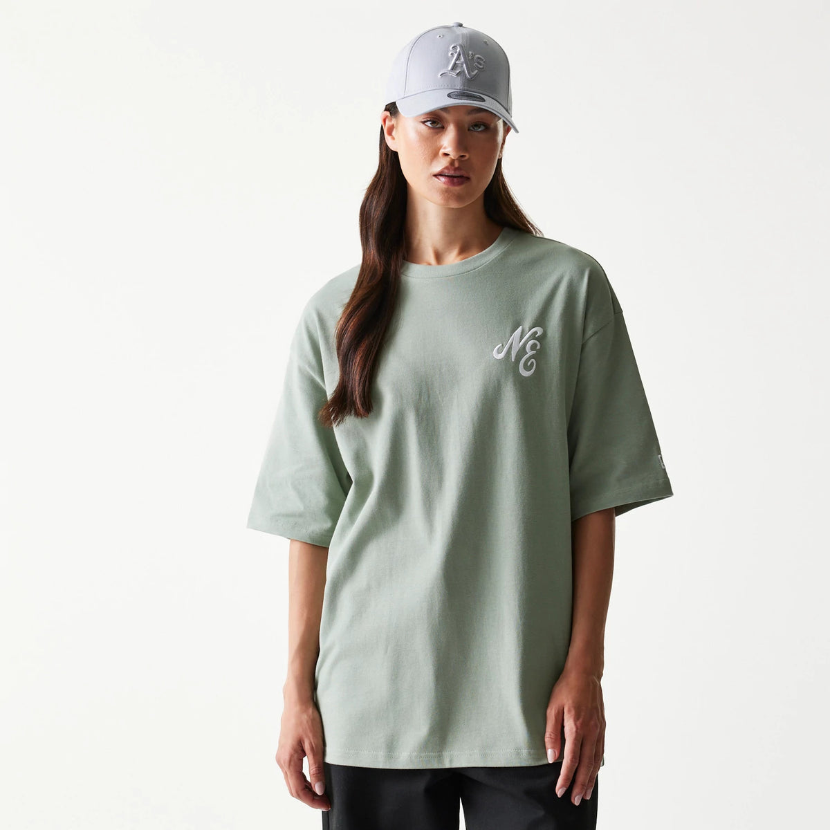New Era Floral Graphic Pastel Green Oversized T-Shirt, front view showing the pastel green colour with an embroidered NE logo on the left chest, displayed on a model paired with a cap and black trousers.