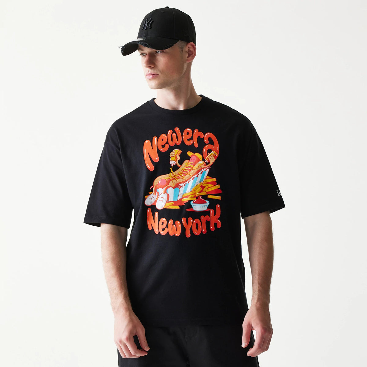 New Era Hotdog Food Graphic Black Oversized T-Shirt, front view showing a colourful hotdog with fries graphic and Newera Newyork lettering on the chest, New Era logo embroidered on the left sleeve, worn by a model with black trousers and cap.