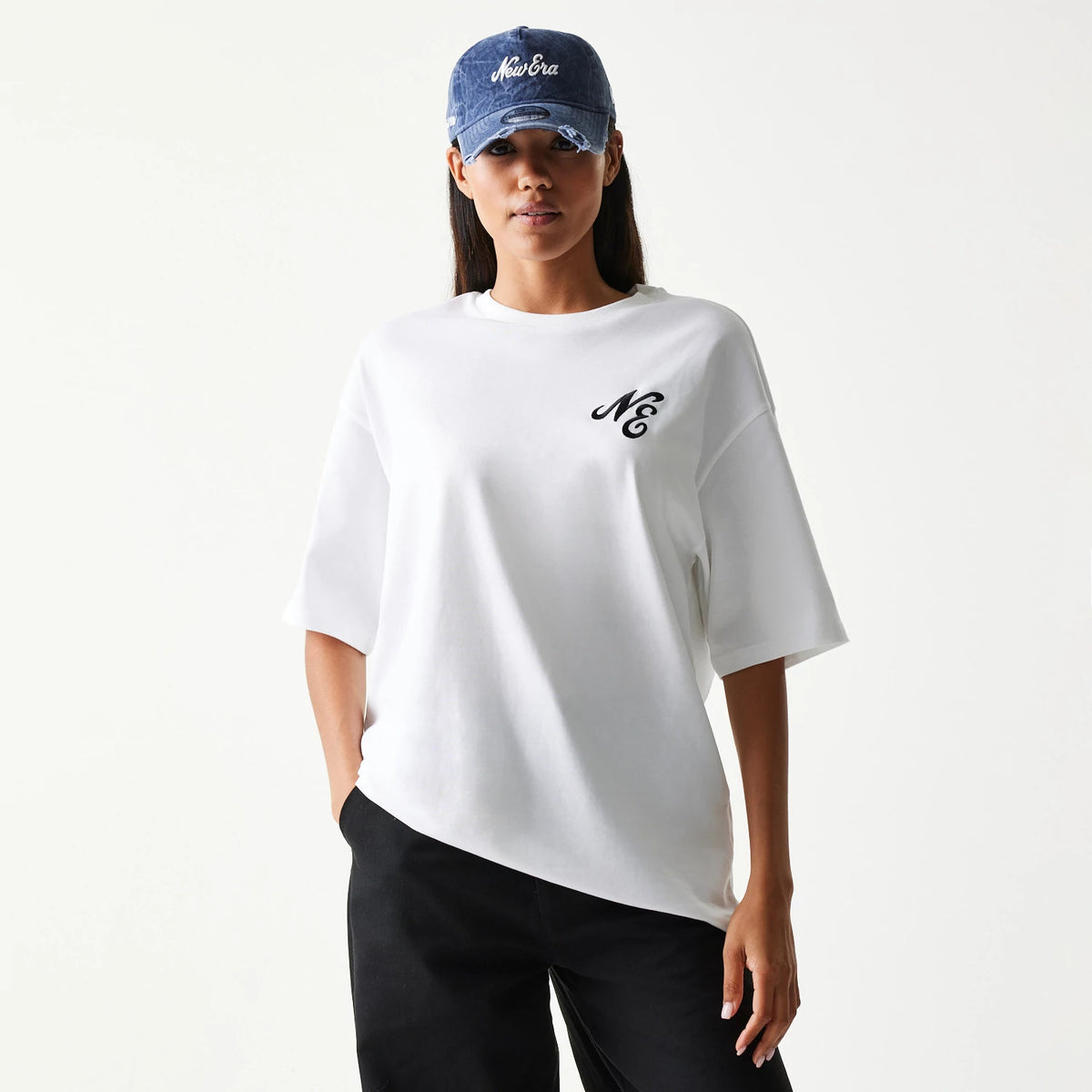 New Era Floral Graphic White Oversized T-Shirt, front view on-model featuring a black embroidered NE logo on the left chest, paired with black trousers and a blue cap with a white embroidered New Era logo on the front.