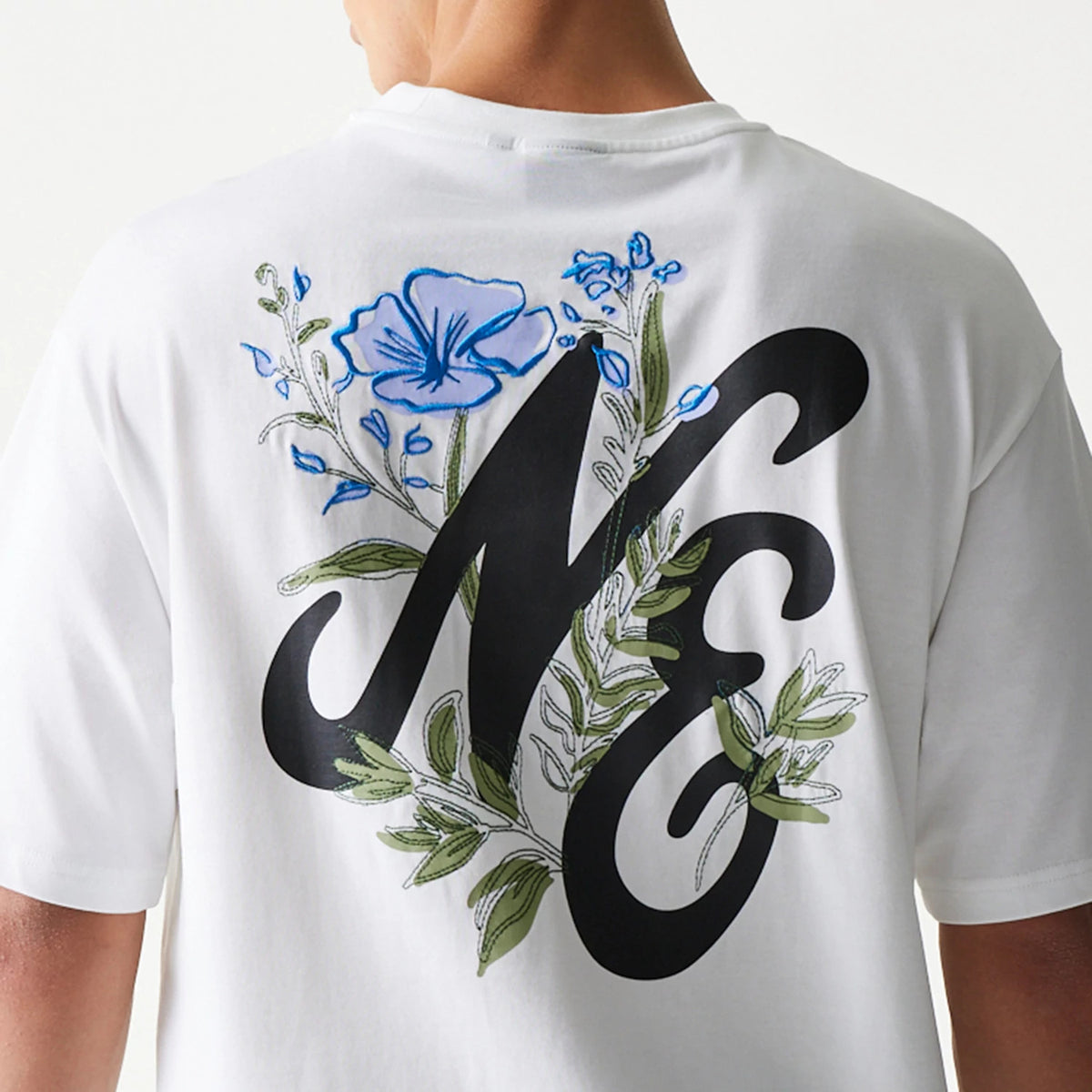 New Era Floral Graphic White Oversized T-Shirt, back view showing a black NE graphic adorned with delicate embroidered blue flowers and green foliage that extend across the upper back, worn by a model.