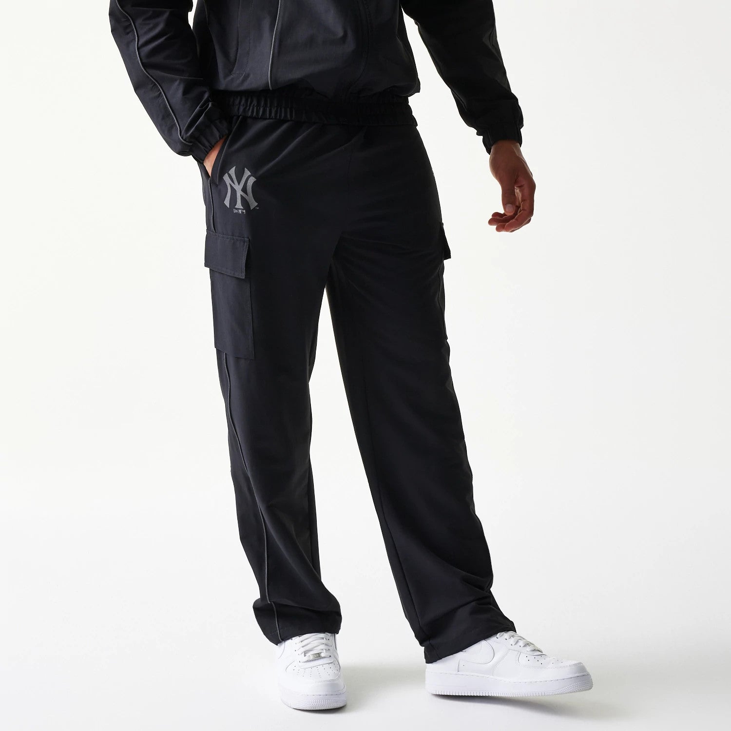 New Era New York Yankees MLB Utility Black Straight Leg Cargo Joggers, front view on a model wearing white trainers and a matching jacket, with the grey Yankees logo on the left thigh and cargo pockets on each side.