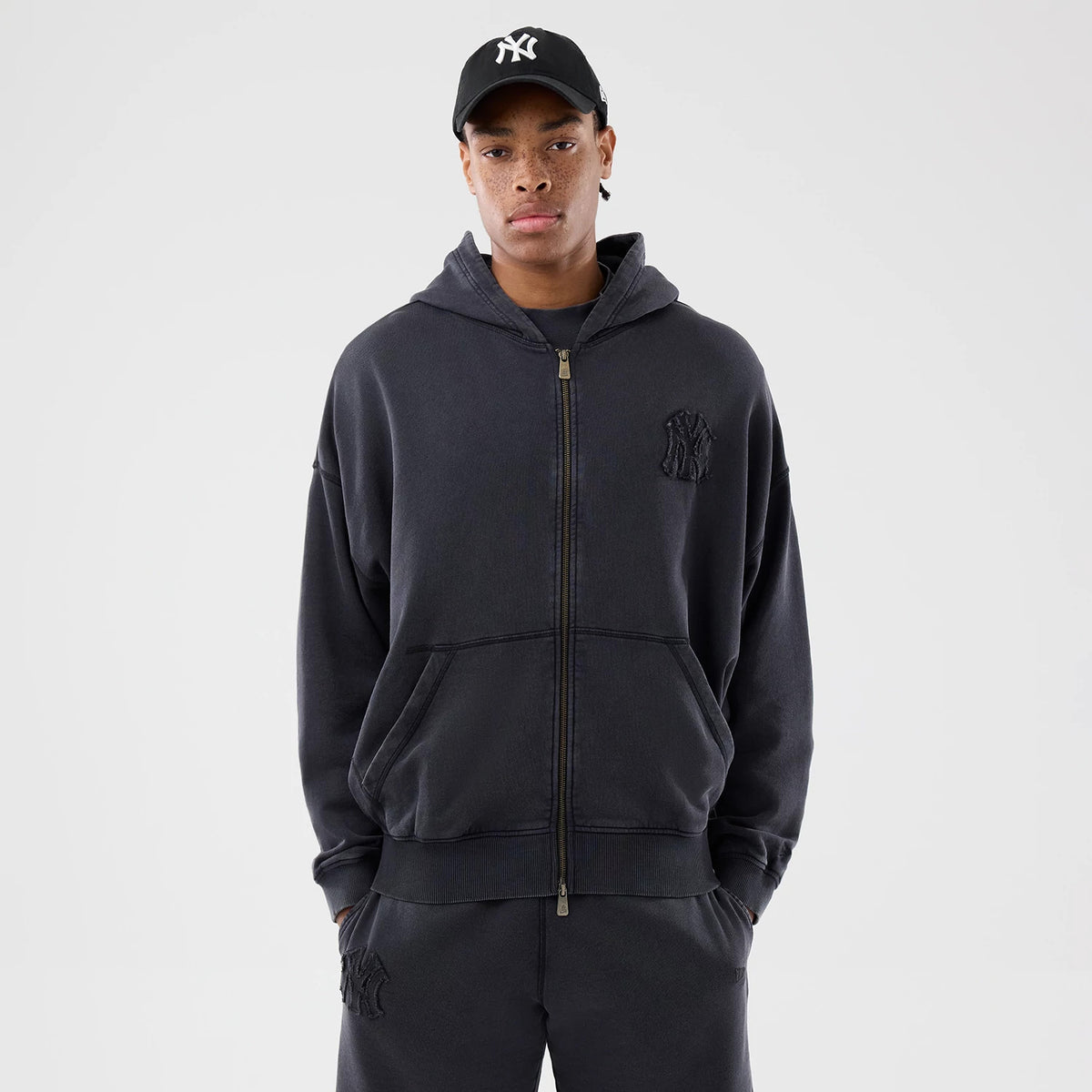 New Era New York Yankees MLB Washed Black Full Zip Hoodie, front view on-model showing the full-length zip, kangaroo pocket, and raised NY logo on the left chest, styled with matching joggers and a black cap featuring an embroidered white NY logo.