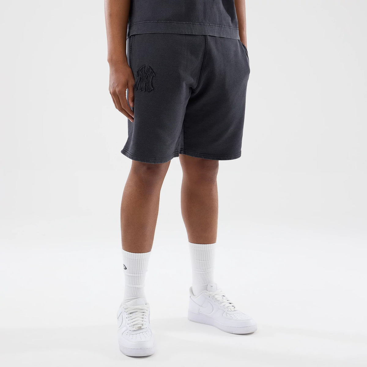 New Era New York Yankees MLB Washed Black Shorts, front view on-model showing the knee-length cut with an embroidered team logo on the left leg near the pocket, styled with white socks and trainers against a neutral background.