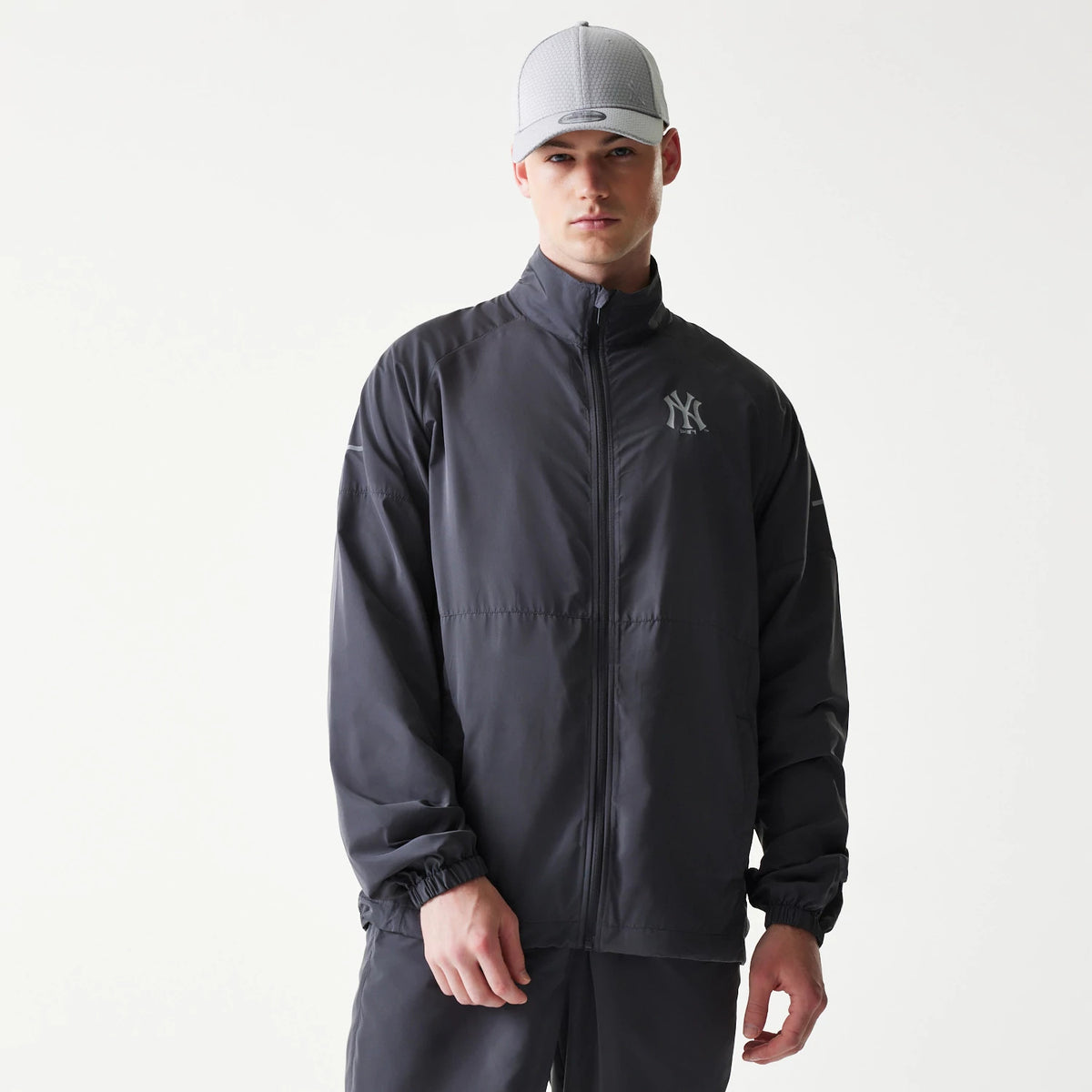 New Era New York Yankees MLB Dark Grey Jacket, front view on a model showing the full-length zip, elasticated cuffs, and a raised New York Yankees logo on the left chest, paired with dark grey trousers and a light grey cap.