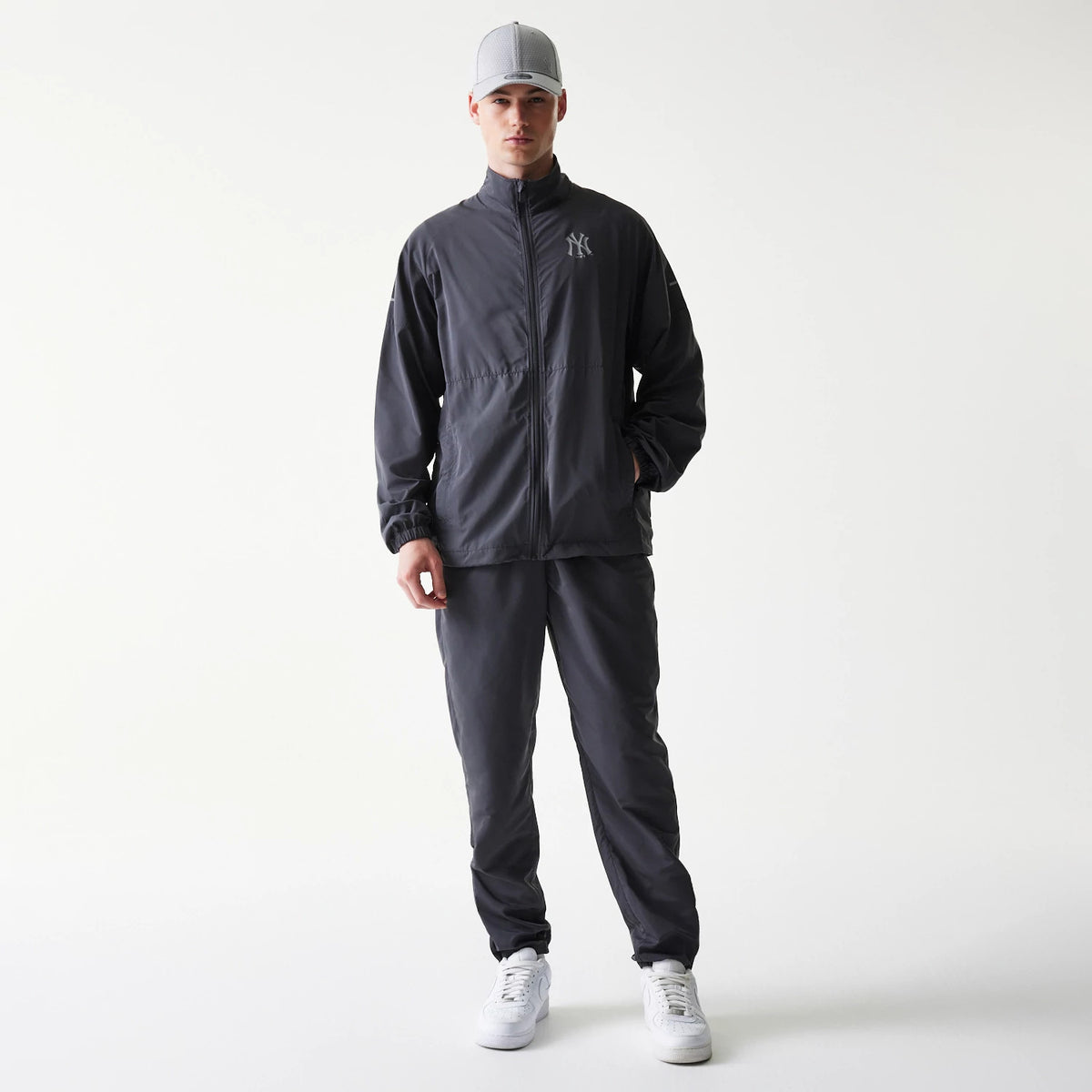New Era New York Yankees MLB Dark Grey Jacket, front view on-model showing a dark grey finish with a raised New York Yankees logo on the left chest, complemented by a central zip closure, styled with matching trousers and a grey cap.