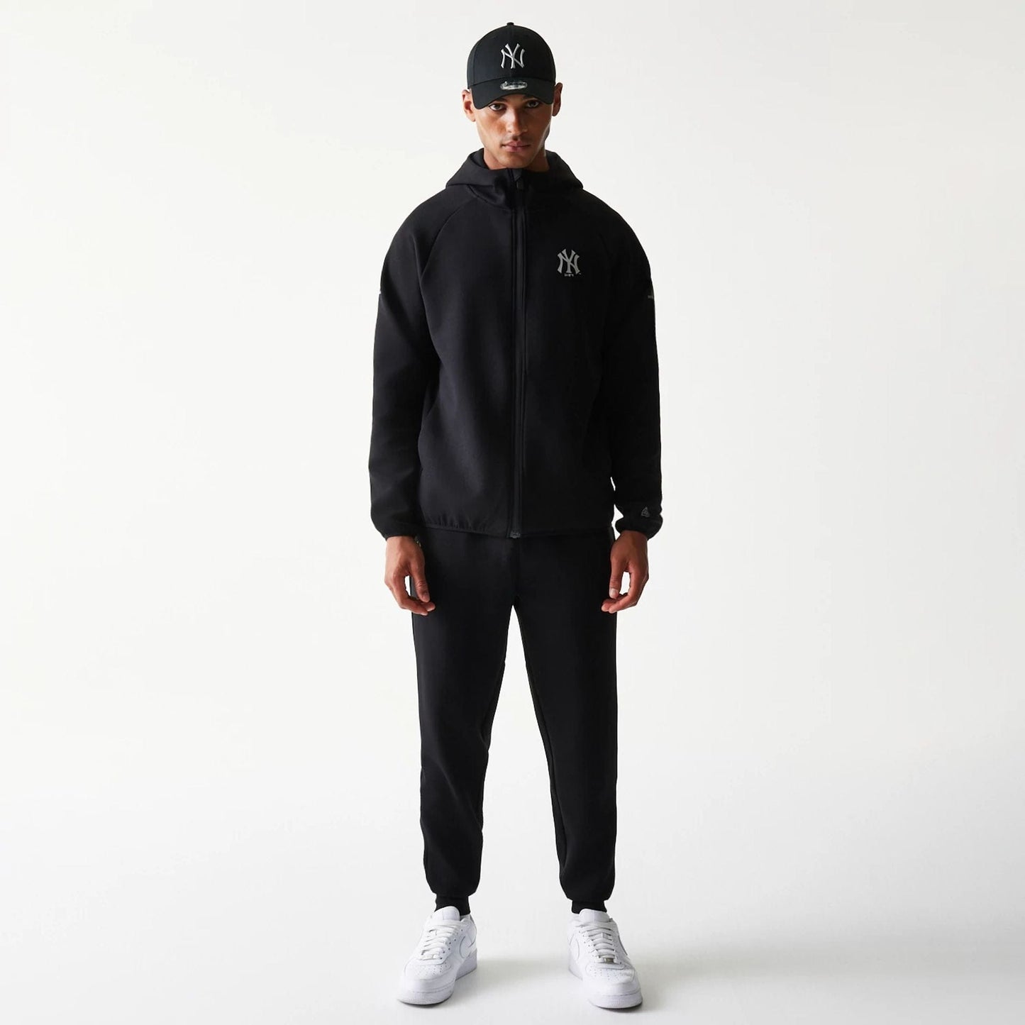 New Era New York Yankees MLB Black Full Zip Hoodie, front view on-model showing the hooded black jacket with an embroidered Yankees logo on the chest, styled with matching black trousers and a black cap adorned with the Yankees logo.