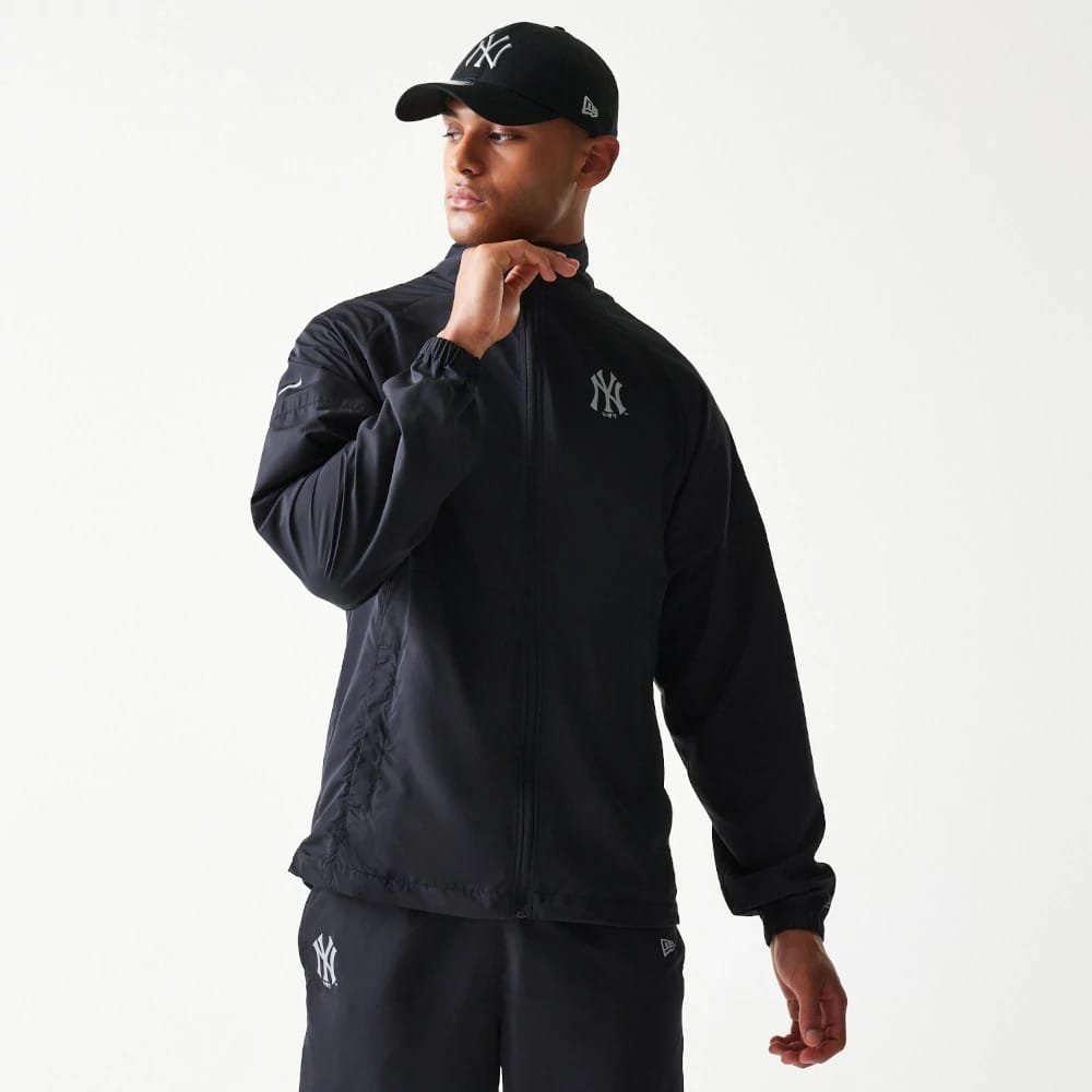 New Era New York Yankees MLB Reflective Strip Black Jacket, front view on a model featuring a printed New York Yankees logo on the left chest, a full zip closure, and elasticated cuffs, paired with matching black trousers and cap.