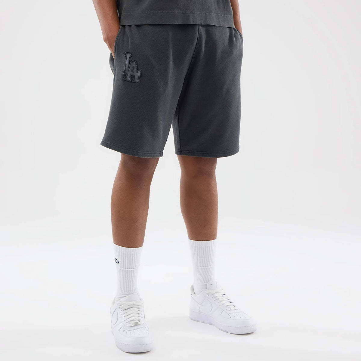 New Era LA Dodgers MLB Washed Dark Grey Shorts, front view showing the dark grey fabric with an embroidered LA logo on the left leg, styled on-model with white socks and trainers, highlighting the relaxed fit and knee-length cut.