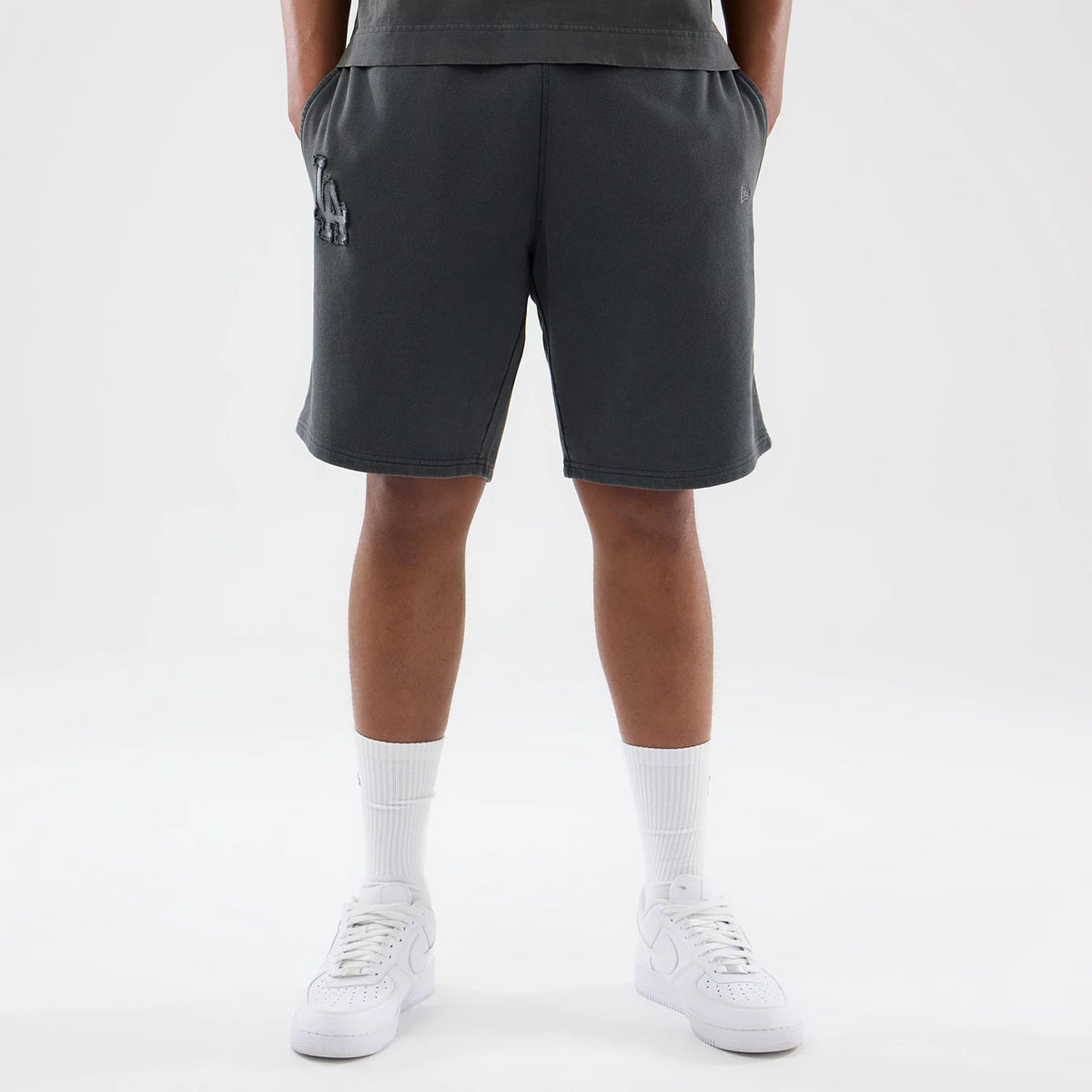 New Era LA Dodgers MLB Washed Dark Grey Shorts, front view showing the dark grey fabric with side pockets, an embroidered LA logo on the left thigh, on-model styled with white trainers and high white socks.
