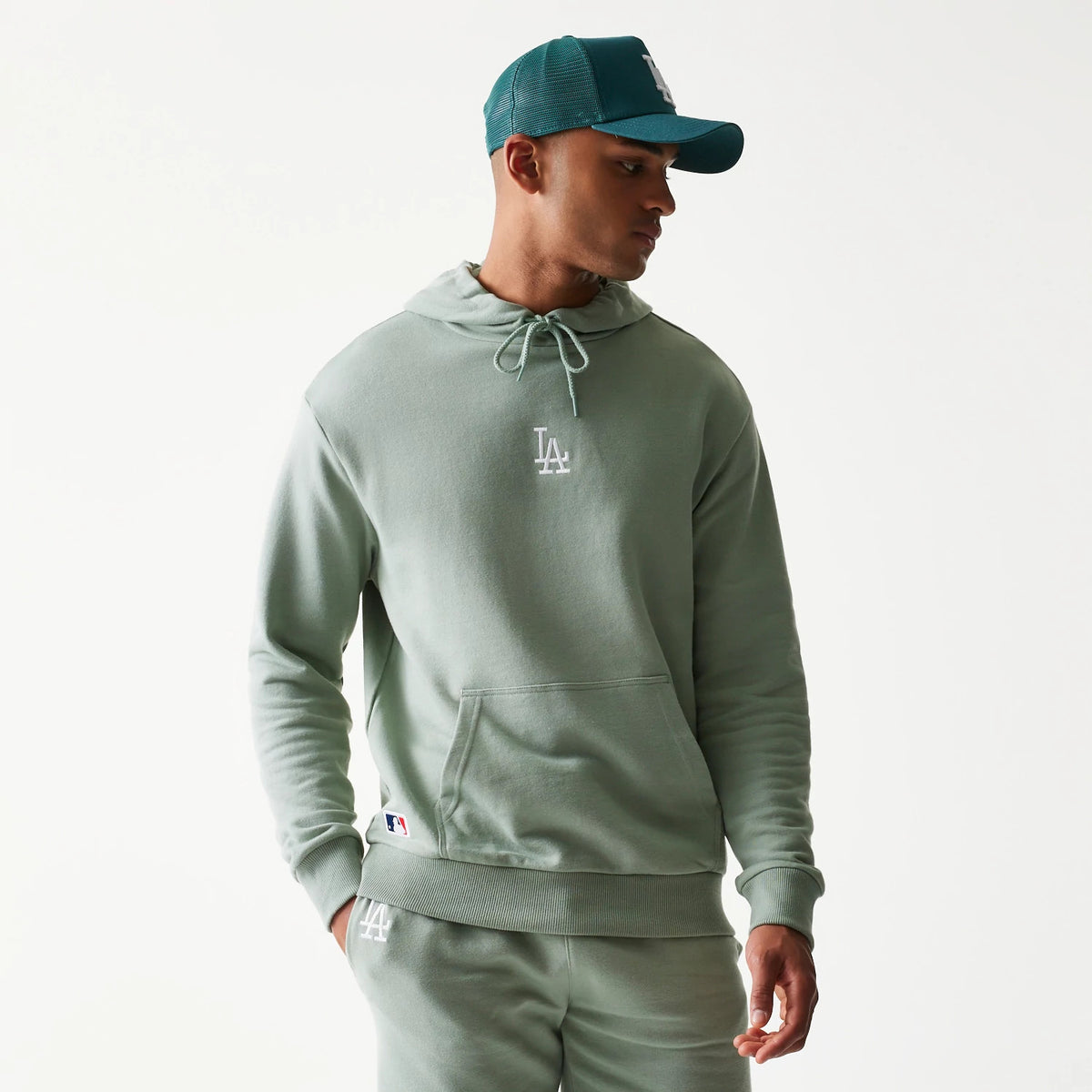 New Era LA Dodgers MLB Midi Green Oversized Pullover Hoodie, front three-quarter view on a model styled with a matching cap, featuring an embroidered white LA logo on the chest and a drawstring hood.