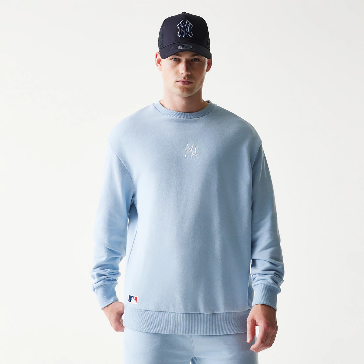 New Era New York Yankees MLB Midi Pastel Blue Oversized Crew Neck Sweater, front view on a model with a black cap, showing the white embroidered Yankees logo on the chest and the MLB logo patch above the ribbed hem.