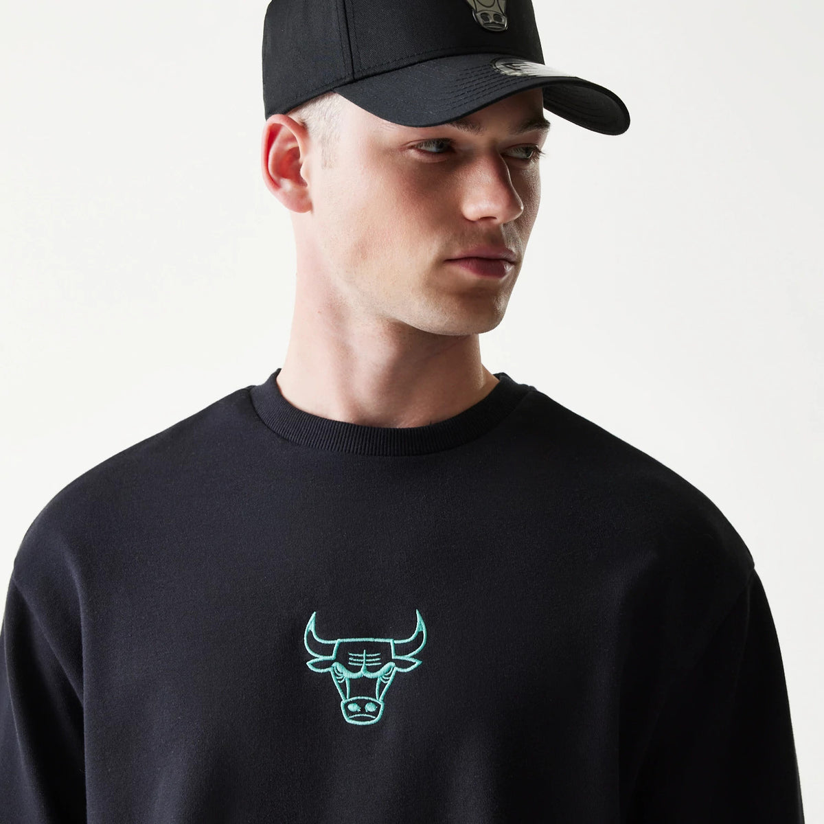 New Era Chicago Bulls NBA Black Oversized Crew Neck Sweater, on-model view featuring the mint green outlined bull head logo embroidered on the chest, paired with a black cap displaying a matching logo on the front panel.