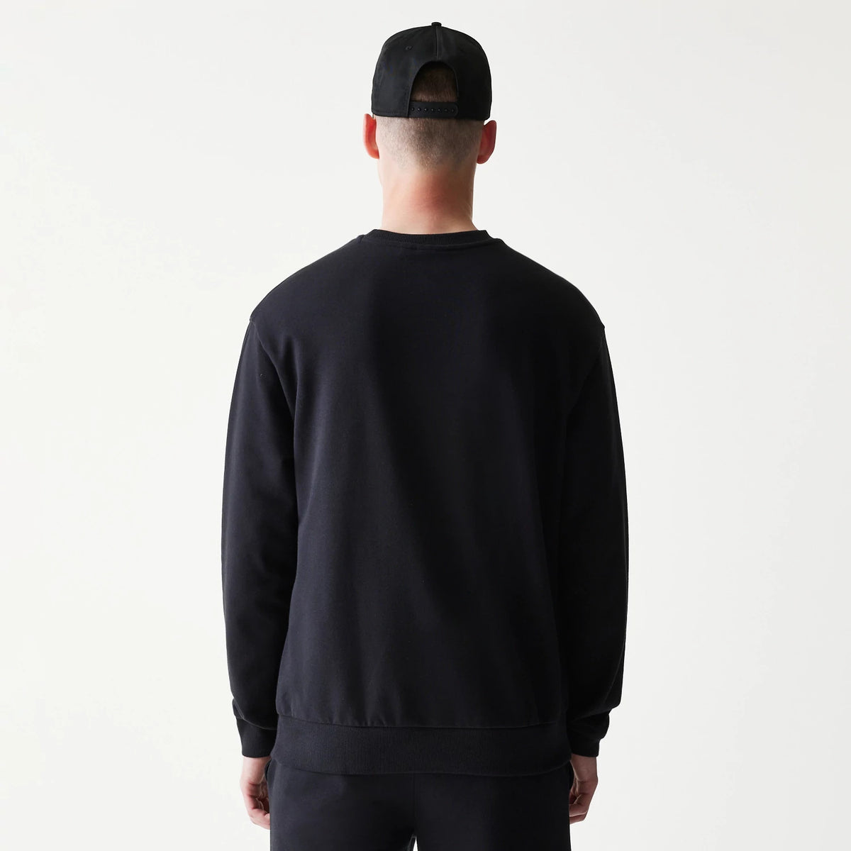 New Era Chicago Bulls NBA Black Oversized Crew Neck Sweater, rear view on a model wearing matching trousers and a black cap, highlighting the plain black back with ribbed cuffs and hem for a classic look.